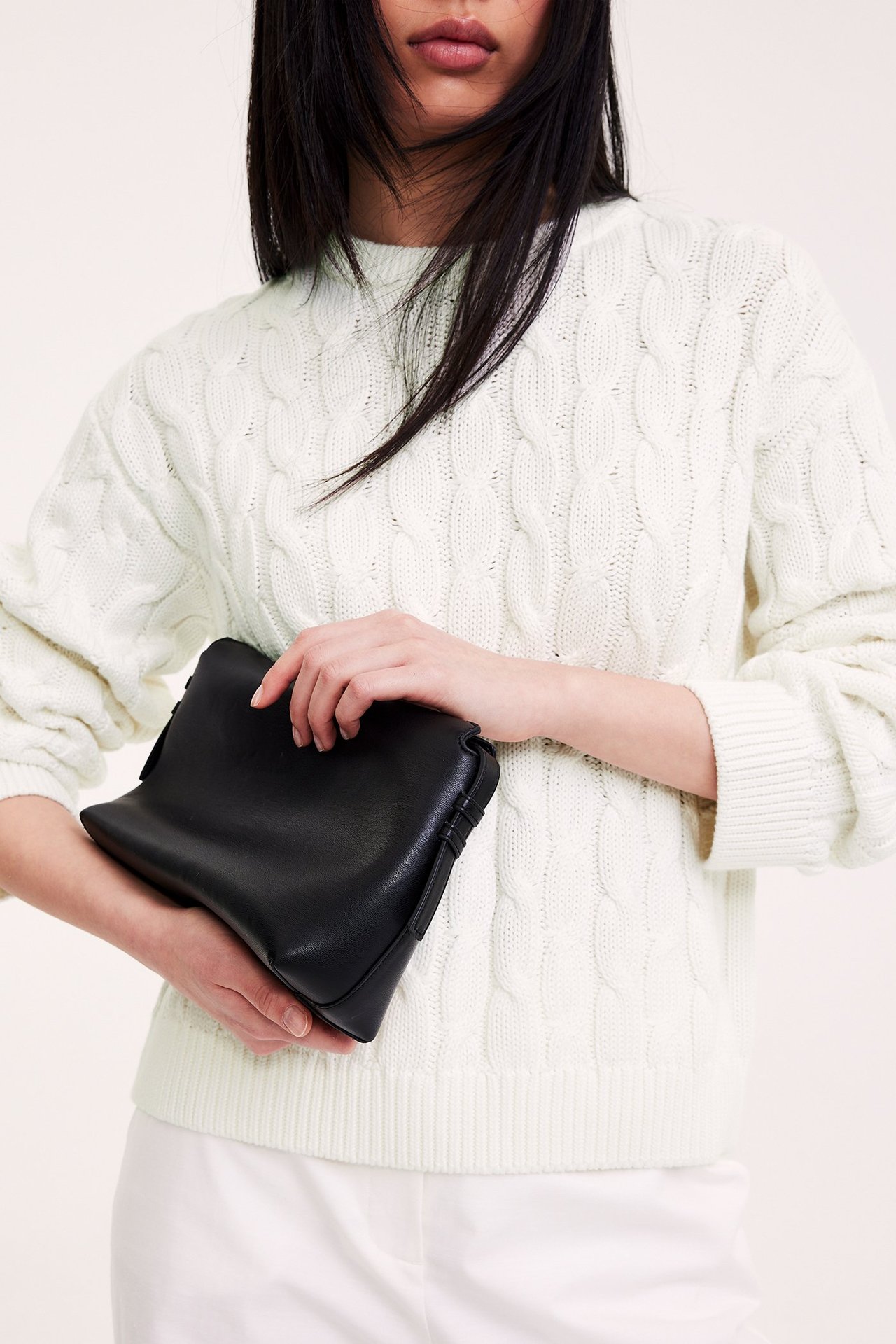 Widok z przodu: kobieta w kremowym swetrze z warkoczowym splotem, trzymająca czarną torebkę.