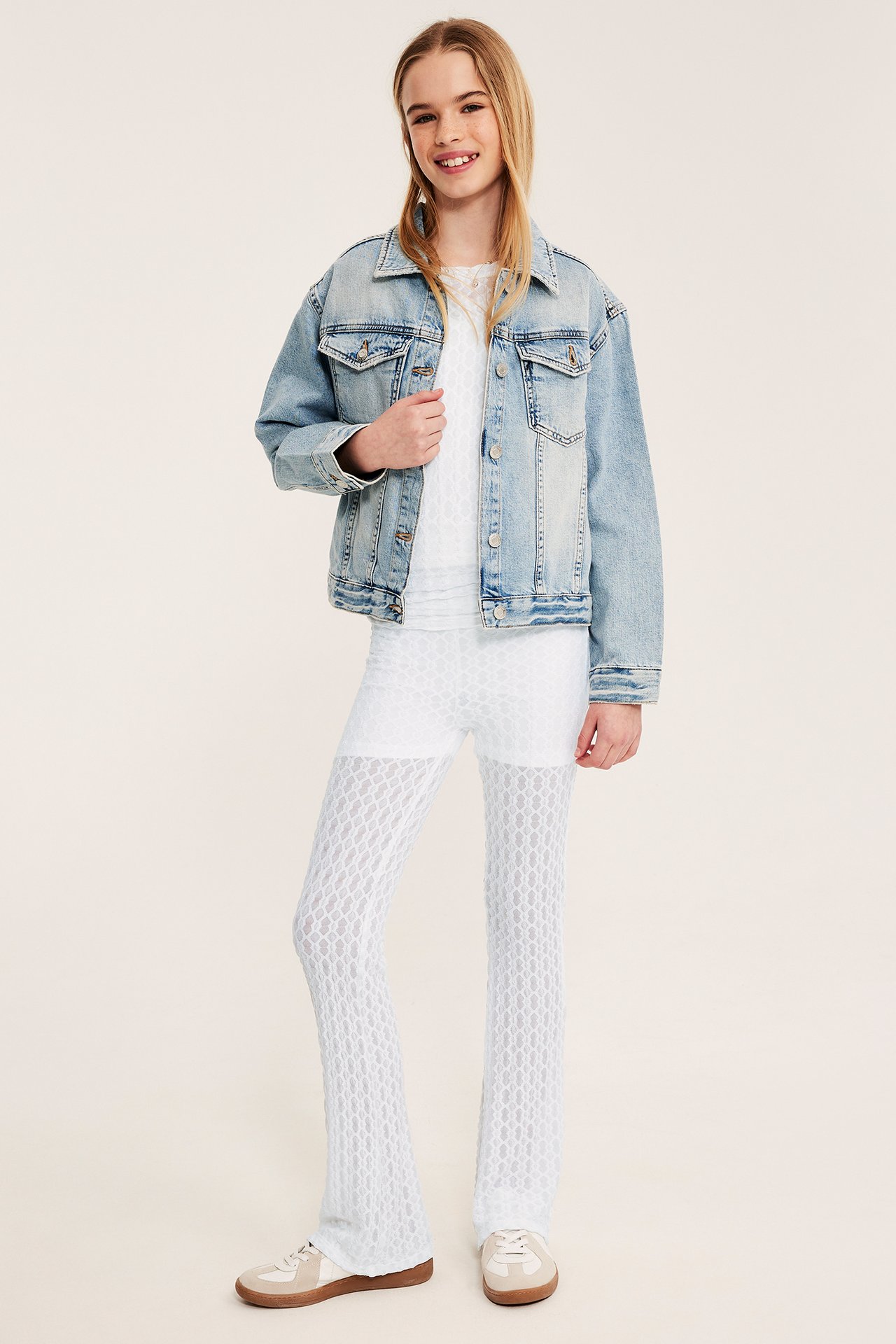 Framifrån: Barn i vita yogabyxor med spetsmönster och utsvängda ben, vit topp, ljusblå jeansjacka, sneakers.