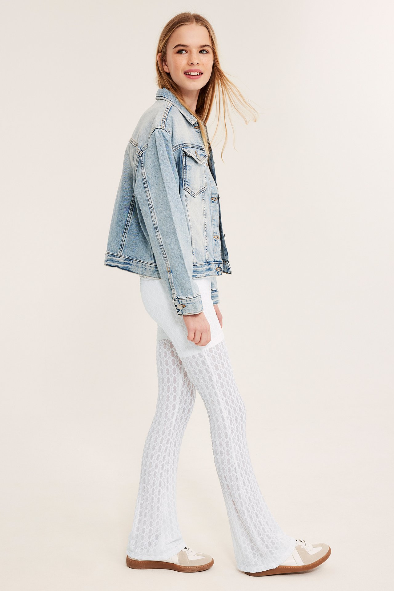 Sidovy av ett leende barn i vita spetsmönstrade utsvängda yogabyxor, en ljusblå jeansjacka och beiga sneakers.