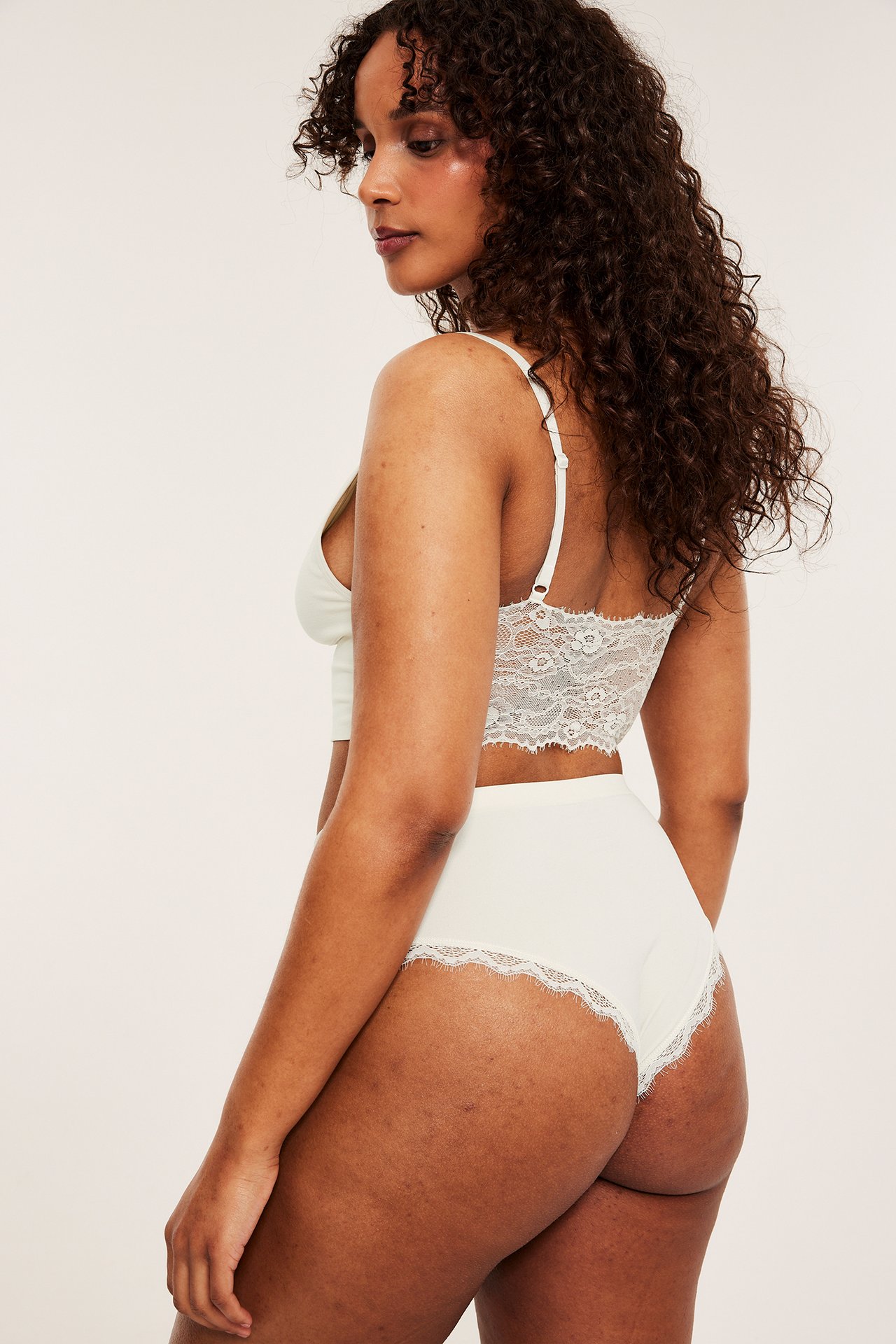 Side-bakfra av en kvinne i off-white cheeky truse med blondekant og matchende bralette.