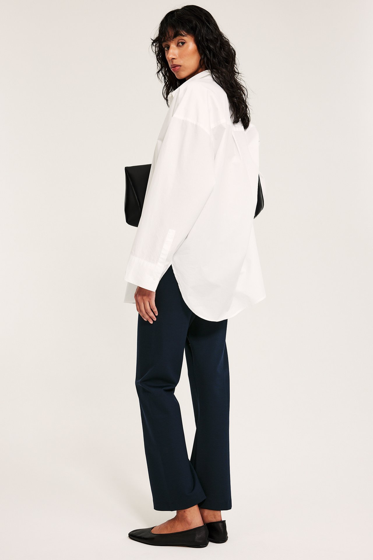 Kobieta w granatowych prostych spodniach pull-on, białej koszuli oversize, czarnych balerinach, trzyma czarną kopertówkę.