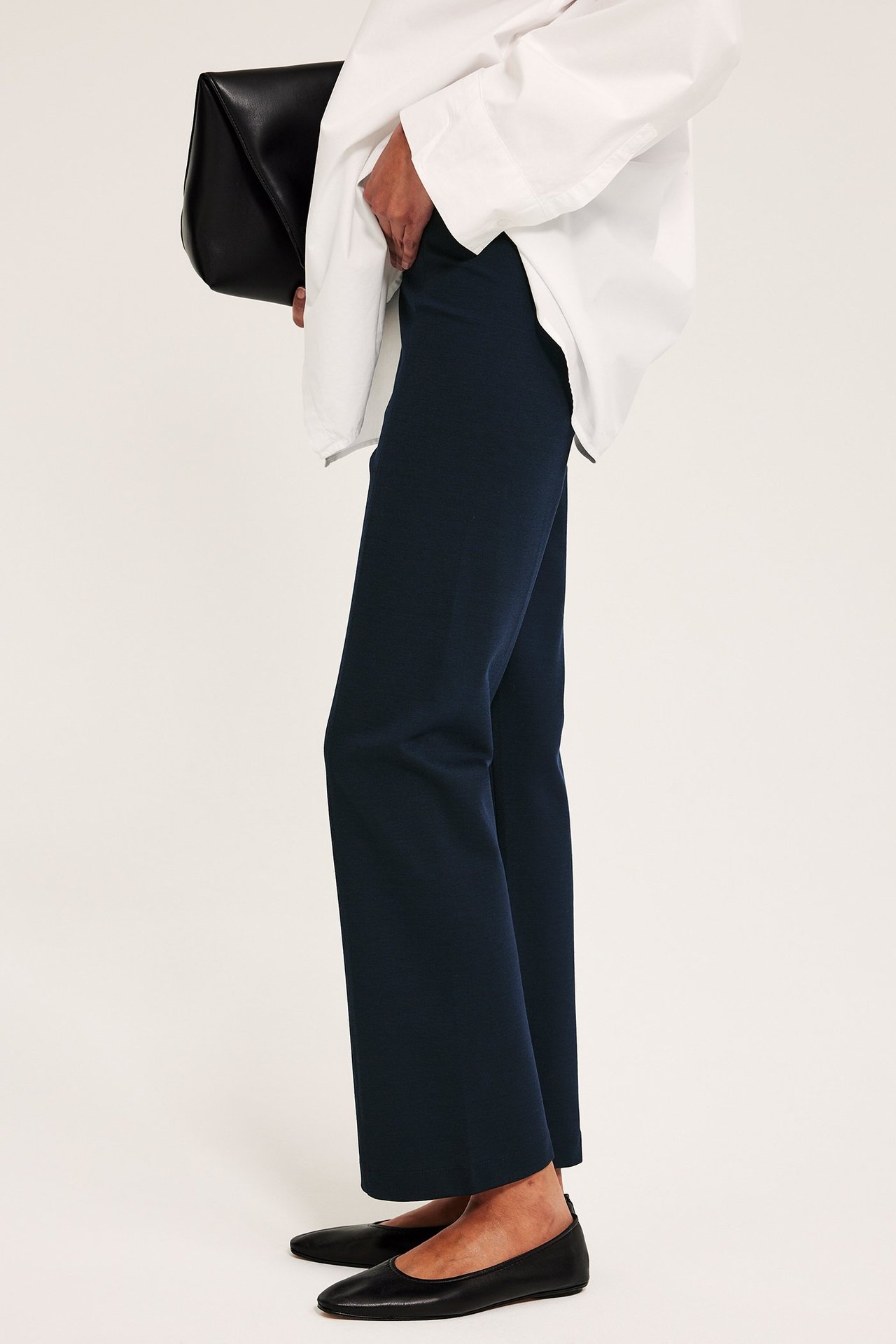 Naisen sivukuva: tummansiniset suorat pull on -housut, valkoinen paita, musta clutch-laukku ja mustat matalat kengät.
