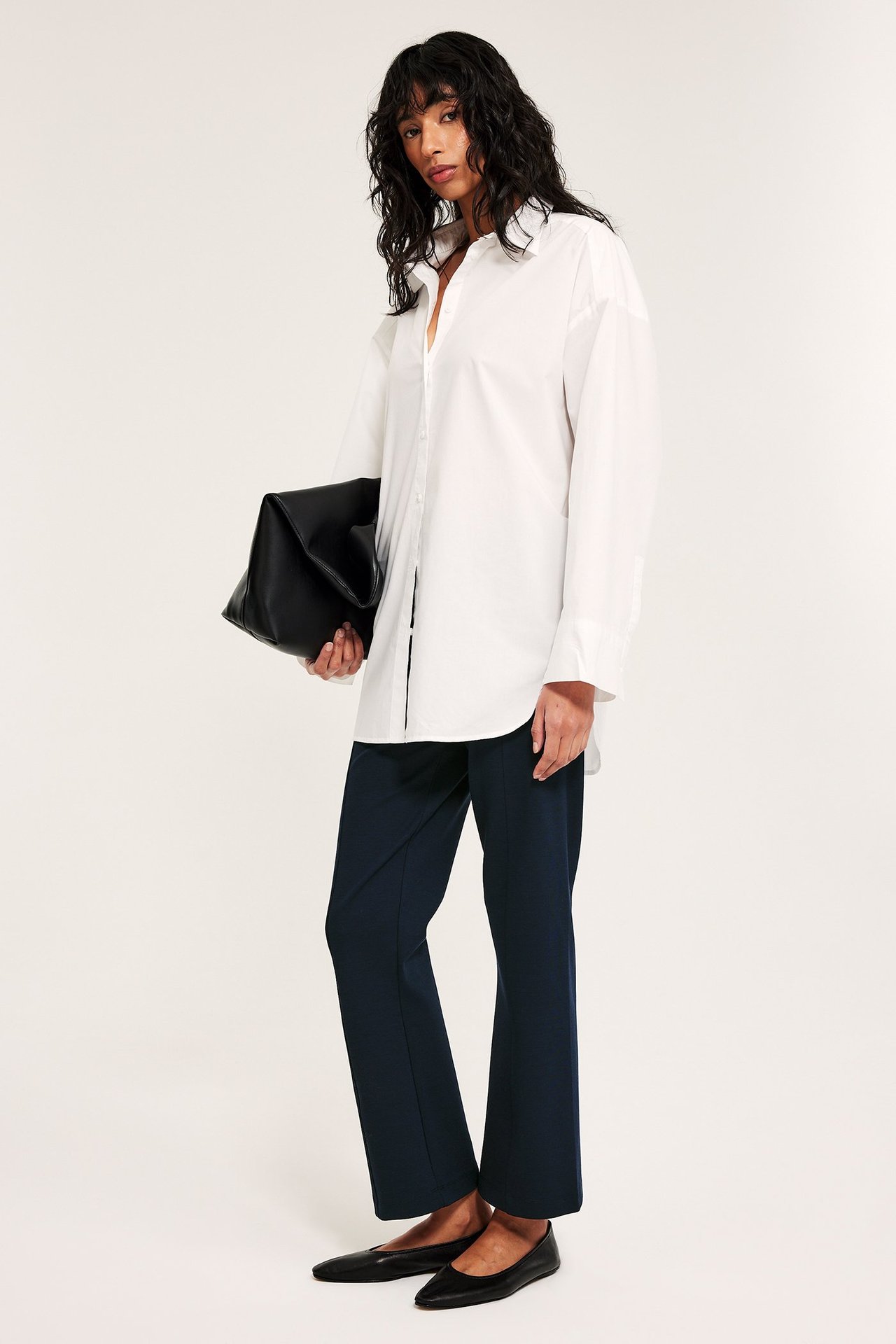 Nainen tummansinisissä suorissa vetohousuissa etusaumoilla, valkoisessa paidassa, mustissa pikkukengissä ja mustassa laukussa.