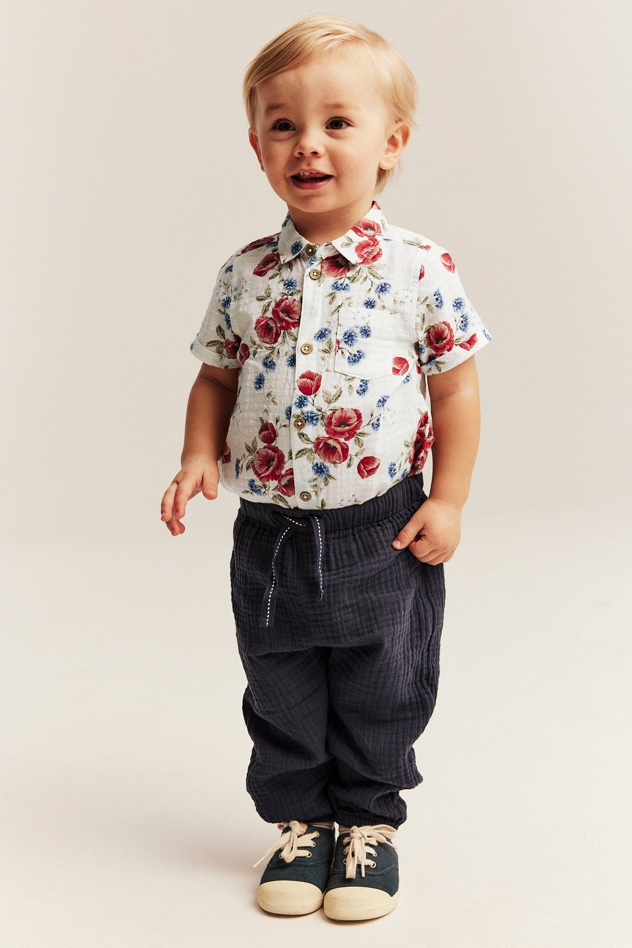 Forfra av en babygutt i en offwhite blomstret kortermet bomulls-skjortebody, mørkeblå bukser og marineblå sko.
