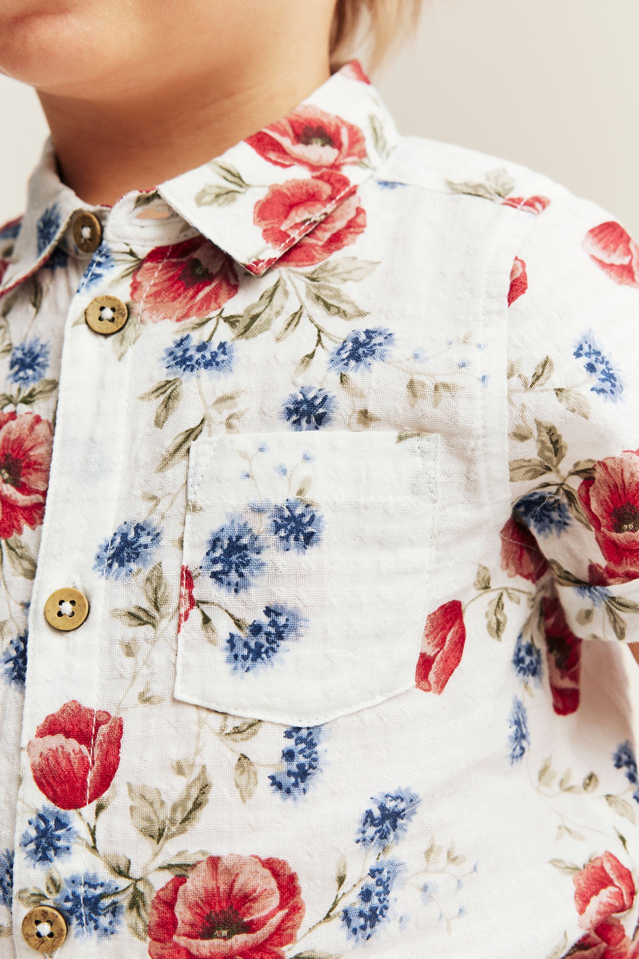 Nærbilde: barn i offwhite blomstret kortermet skjortebody med røde, blå blomster, knapper og brystlomme.