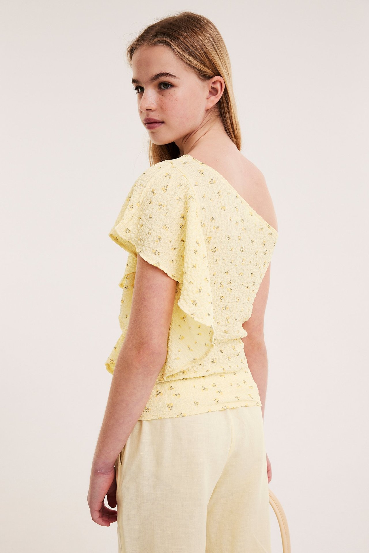 Sidebilde av et barn i lys gul mønstret one shoulder-topp med volanger og lys gule bukser.
