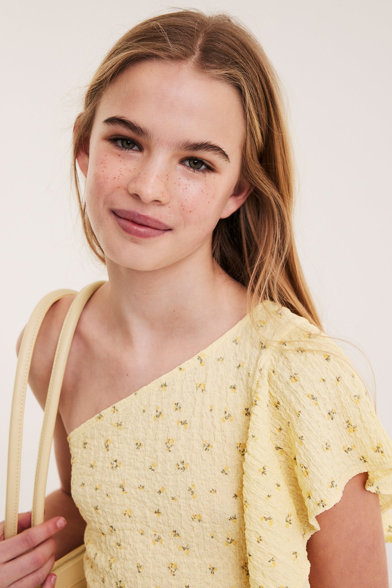 Nærbilde forfra av jente i lys gul en-skulder-topp med blomstermønster og rysjer, holder veske.