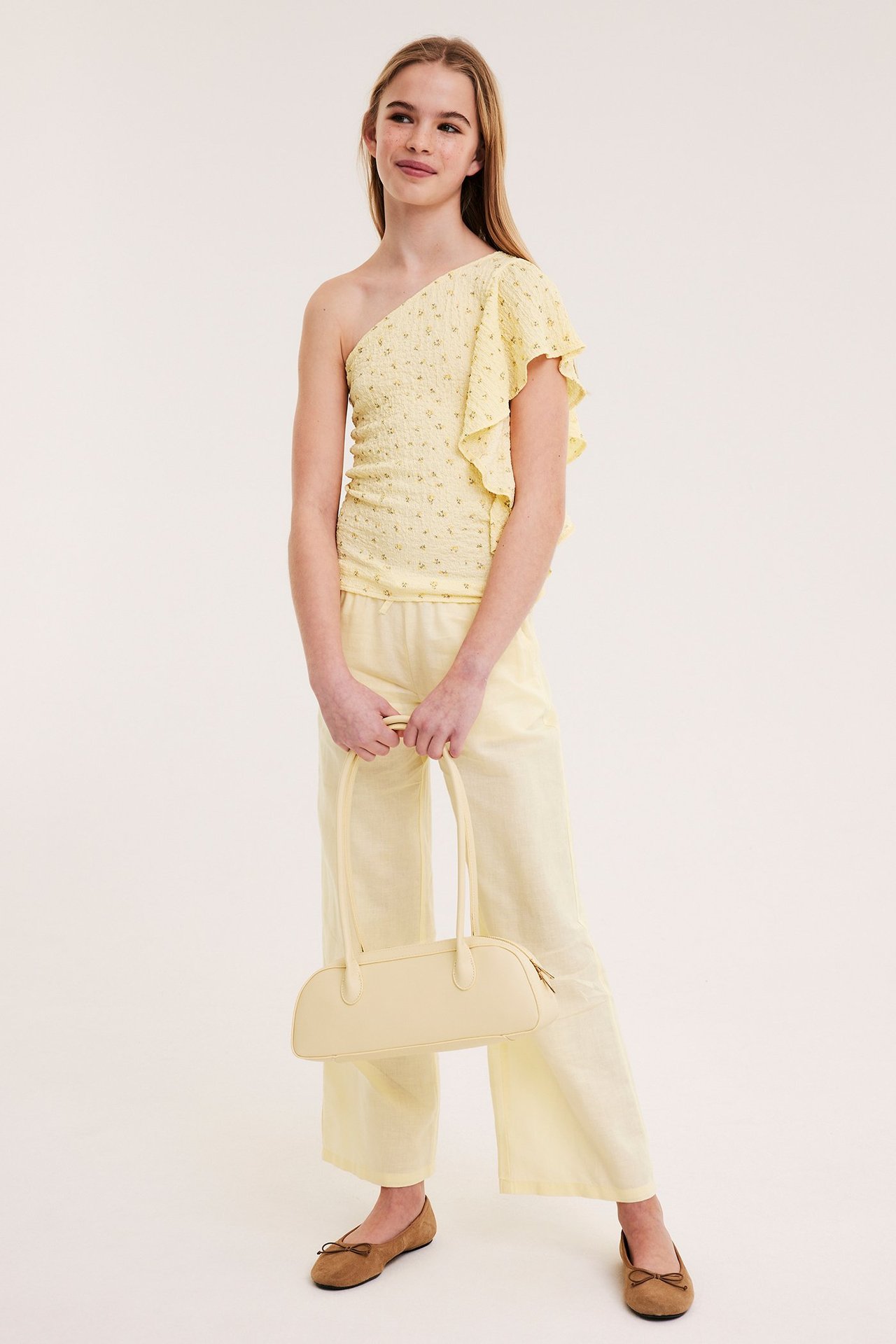 Forfra: Barn iført mønstret gul one-shoulder topp, vide bukser og holder en gul veske.