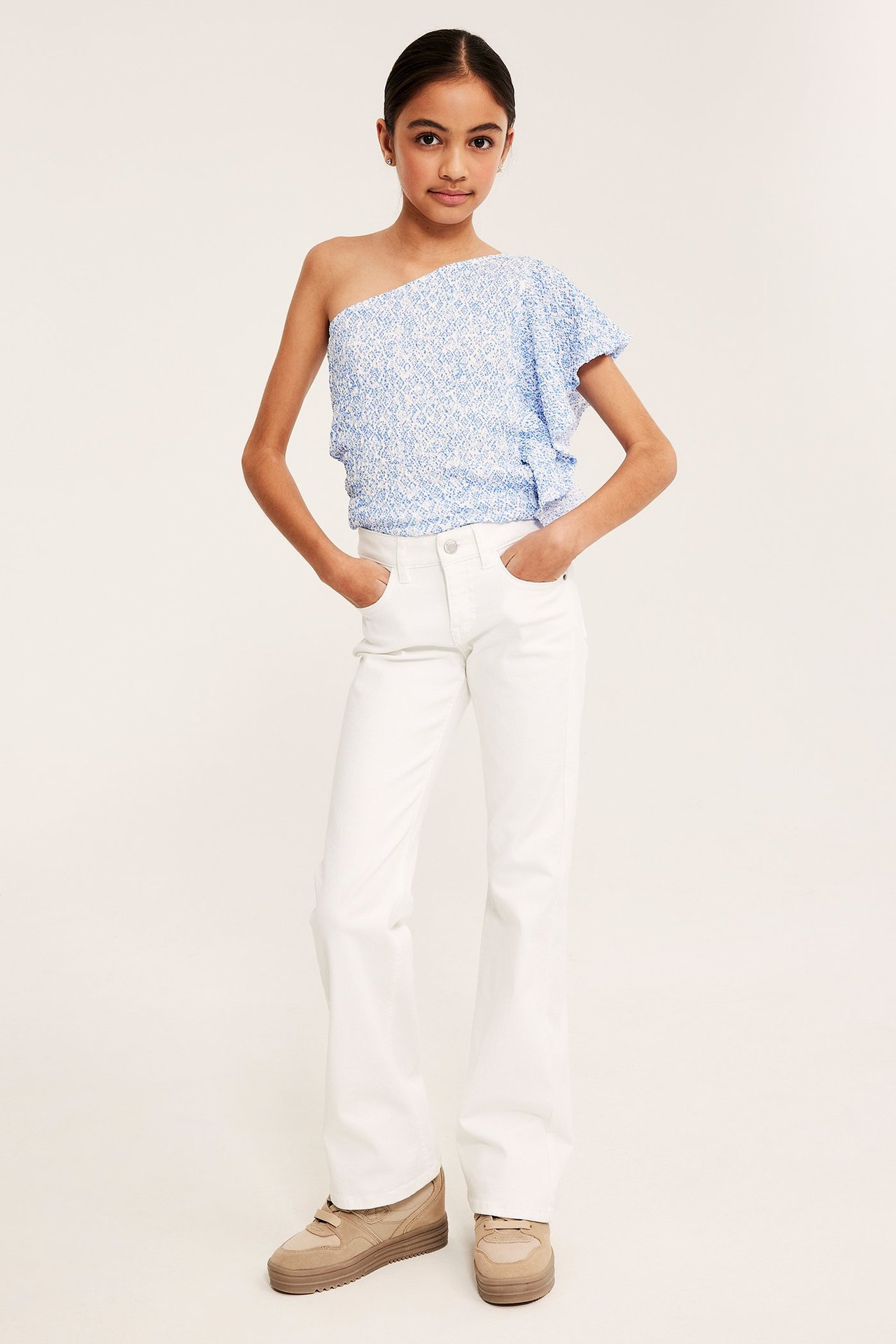 Framifrån, ett barn i blå mönstrad one shoulder-topp med volanger, vita utsvängda jeans och beiga sneakers.