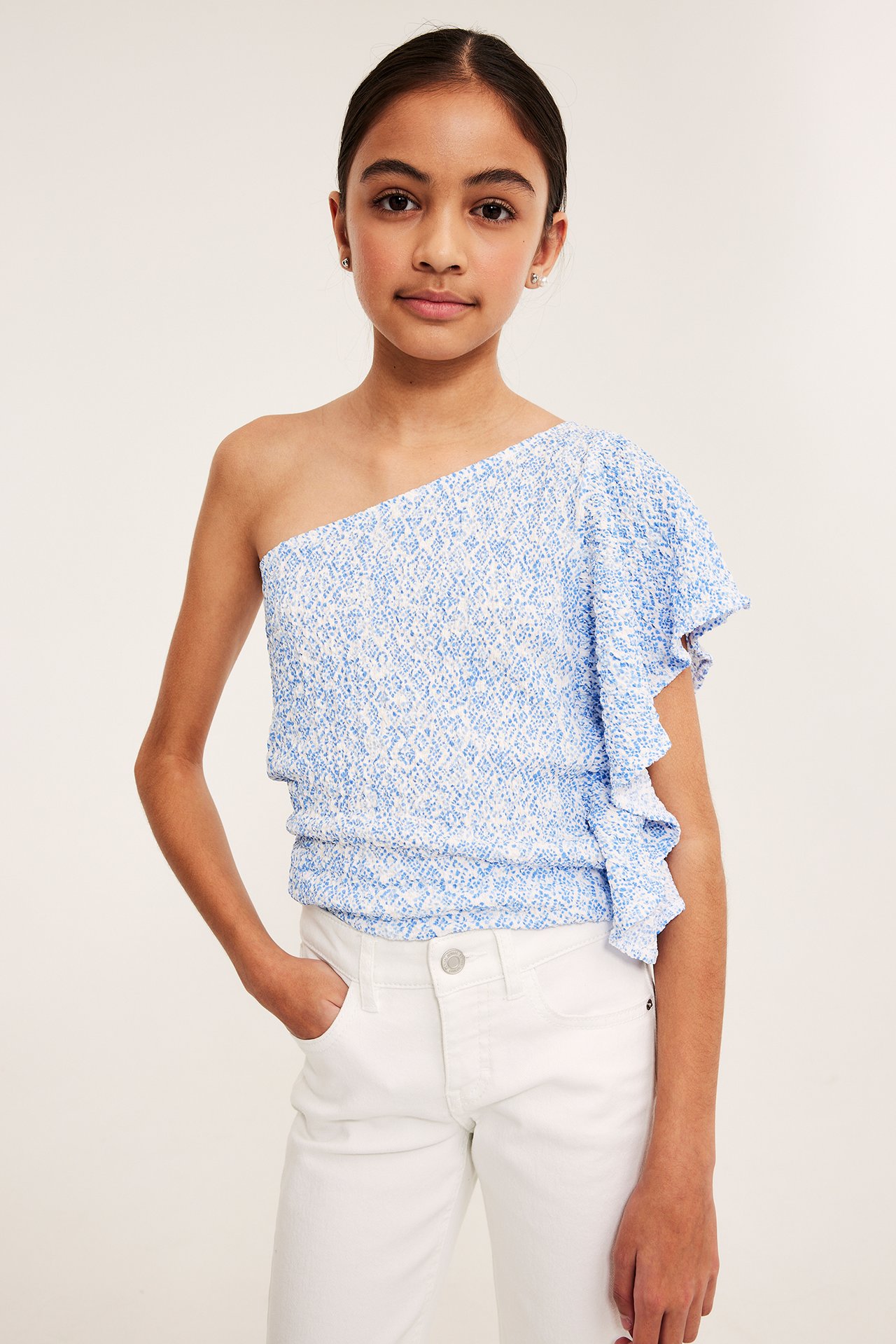 Framifrån, ett barn i en blåmönstrad one shoulder-topp med sidovolang och vita jeans.
