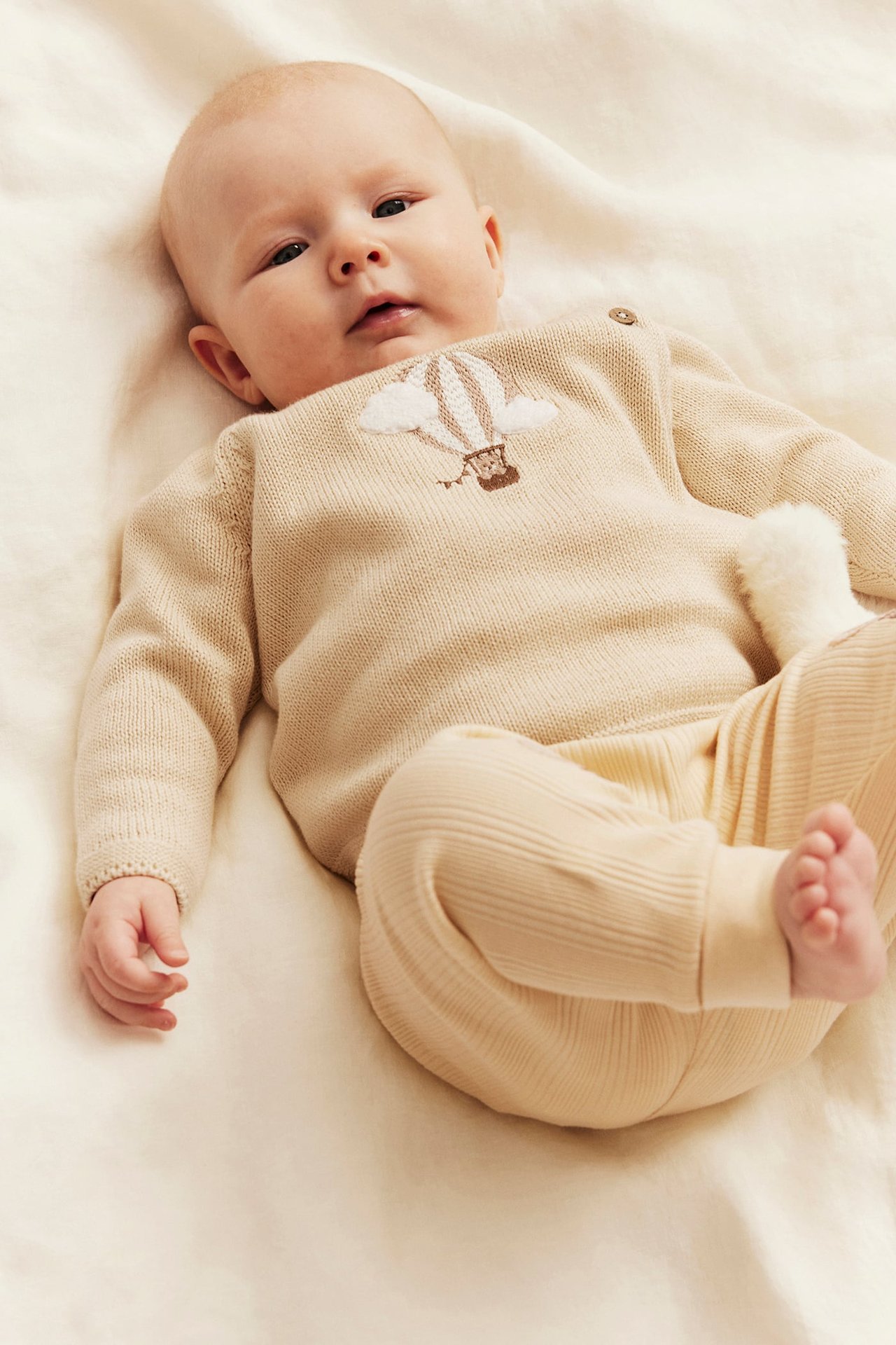 Framifrån, baby i beige stickad tröja med luftballongsbrodyr och matchande ribbstickade leggings.