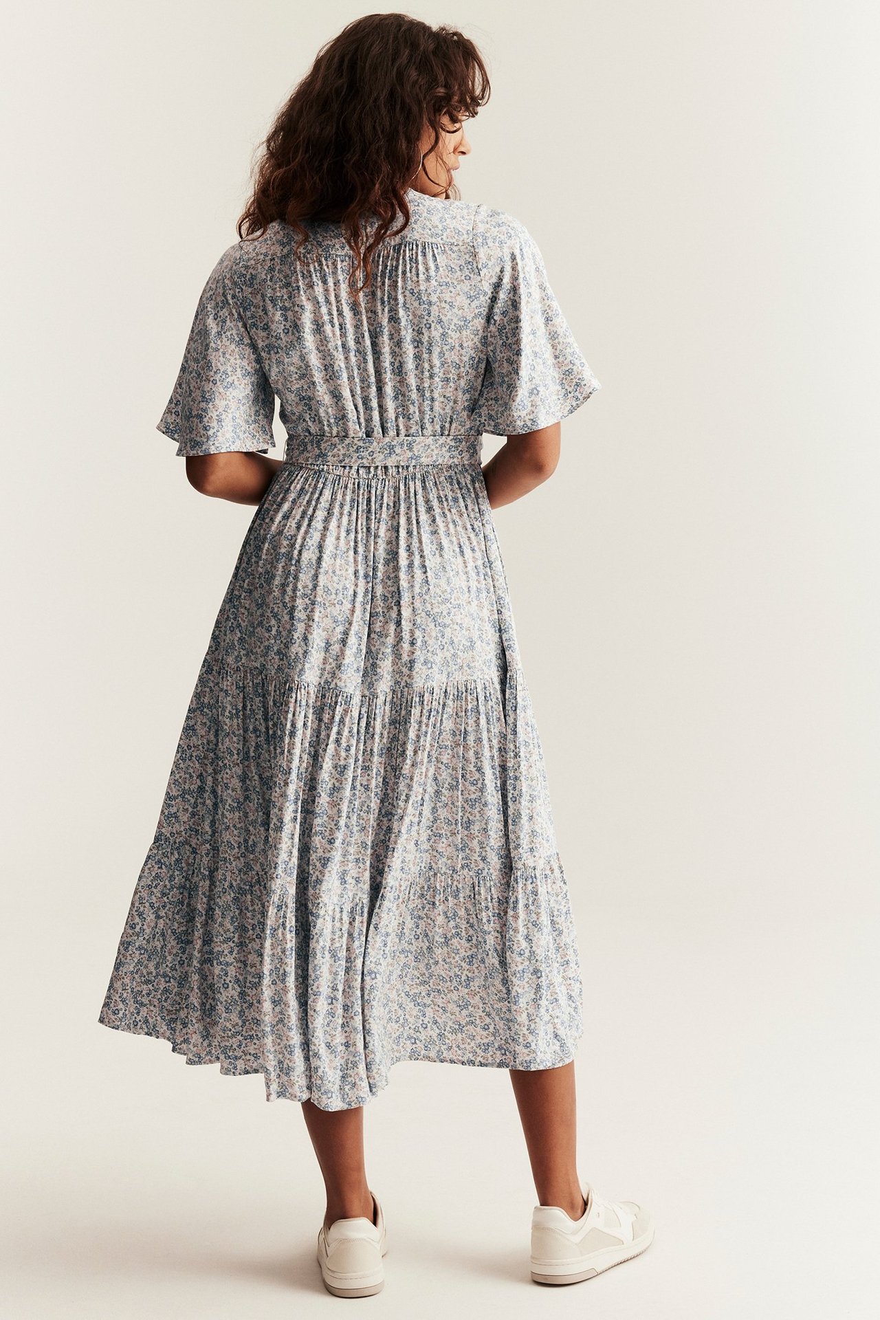 Bakfra: Kvinne i offwhite blomstret viskose-midikjole med korte ermer, knytebelte. Hvite sneakers.