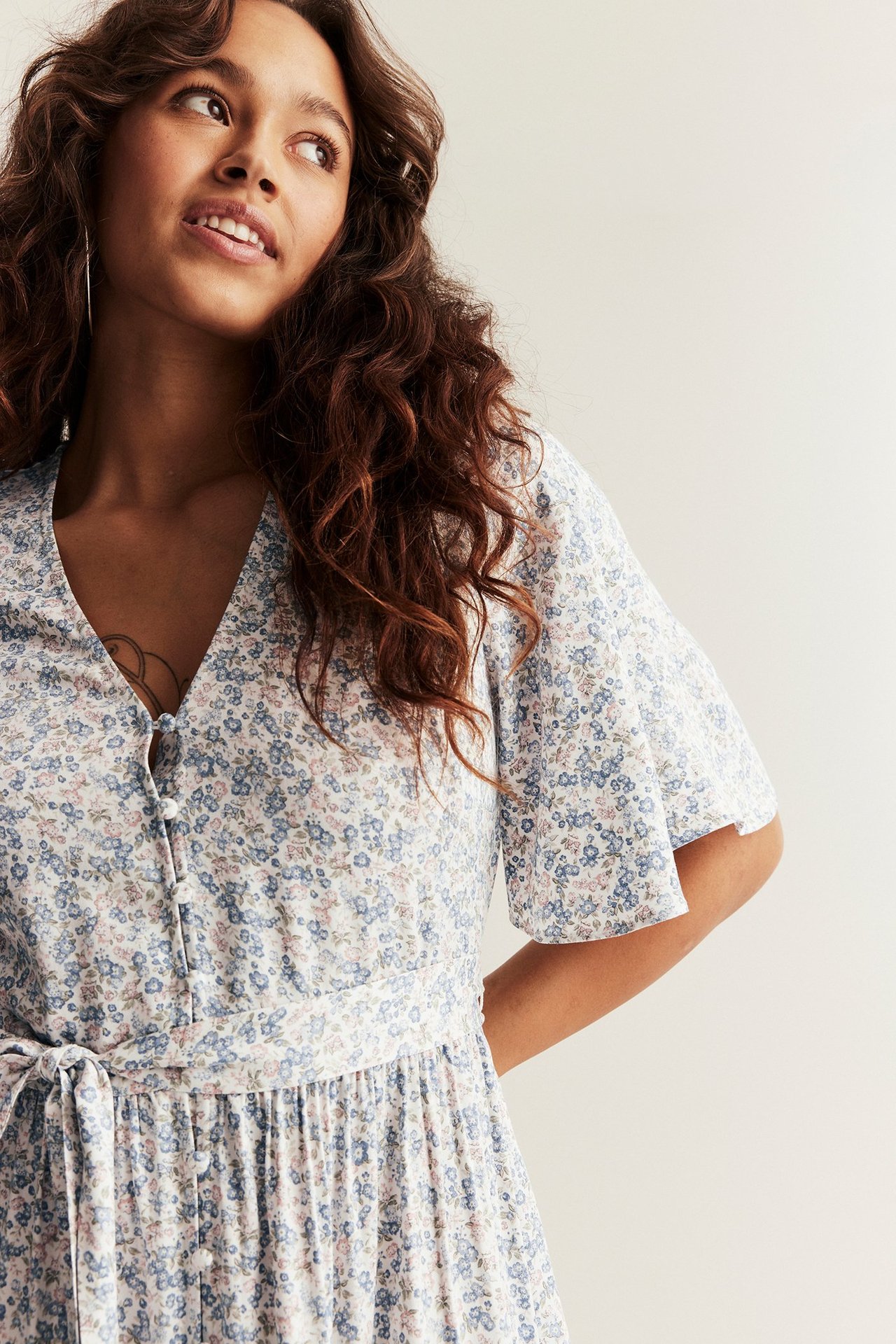 Nærbilde forfra av kvinne i offwhite blomstret viskosekjole med V-hals, korte ermer og knytebelte.