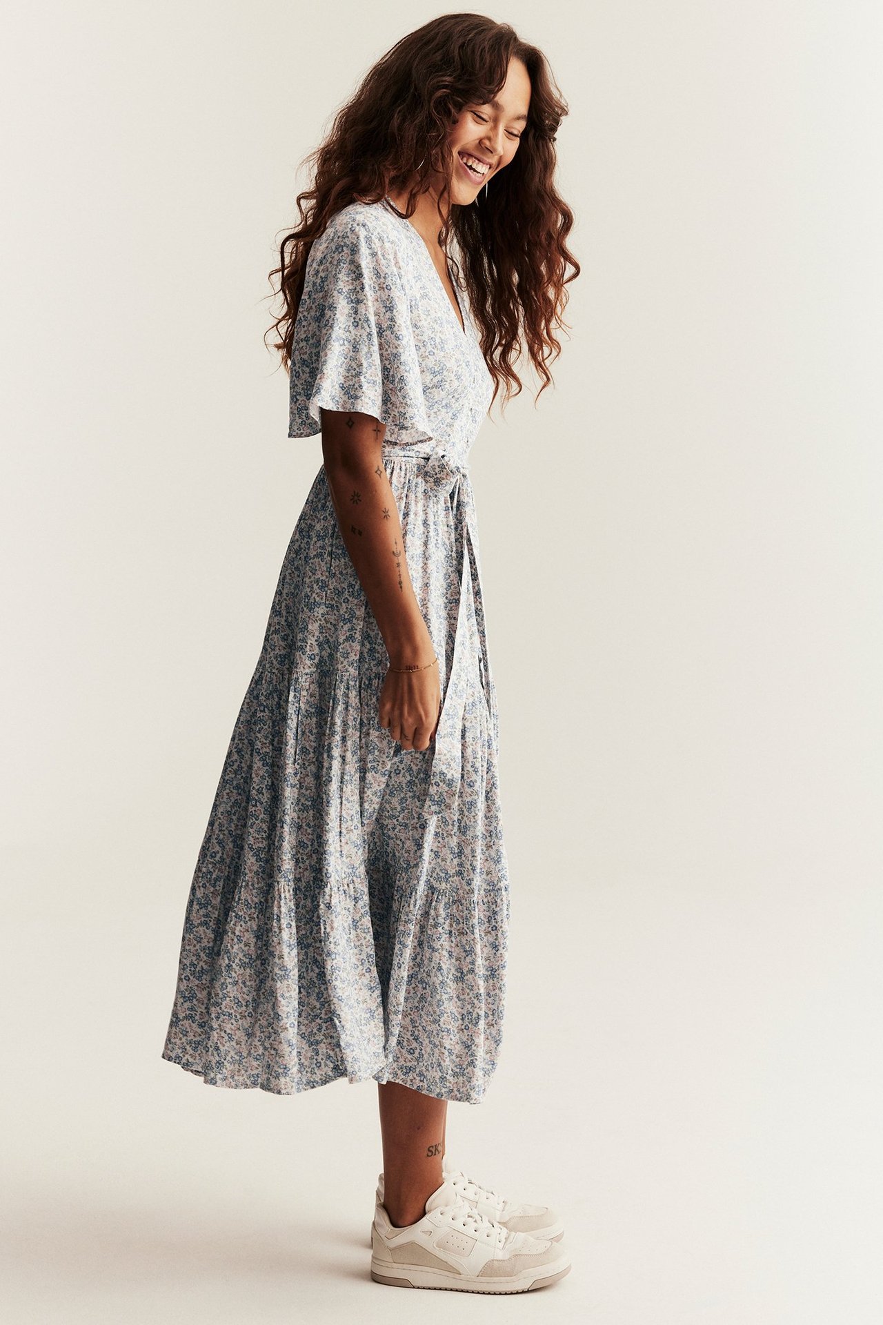 Sidevisning av kvinne i offwhite blomstret viskose-midikjole med korte ermer, knyttet belte og hvite joggesko.