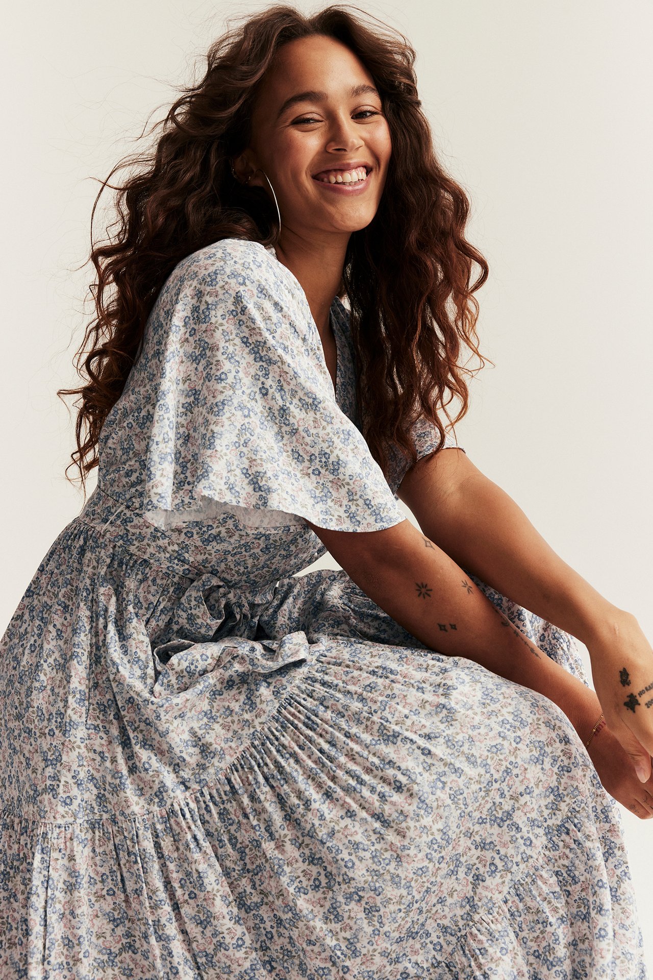 Uśmiechnięta kobieta nosi off-white kwiecistą wiskozową sukienkę z dekoltem V, krótkimi rękawami i paskiem.