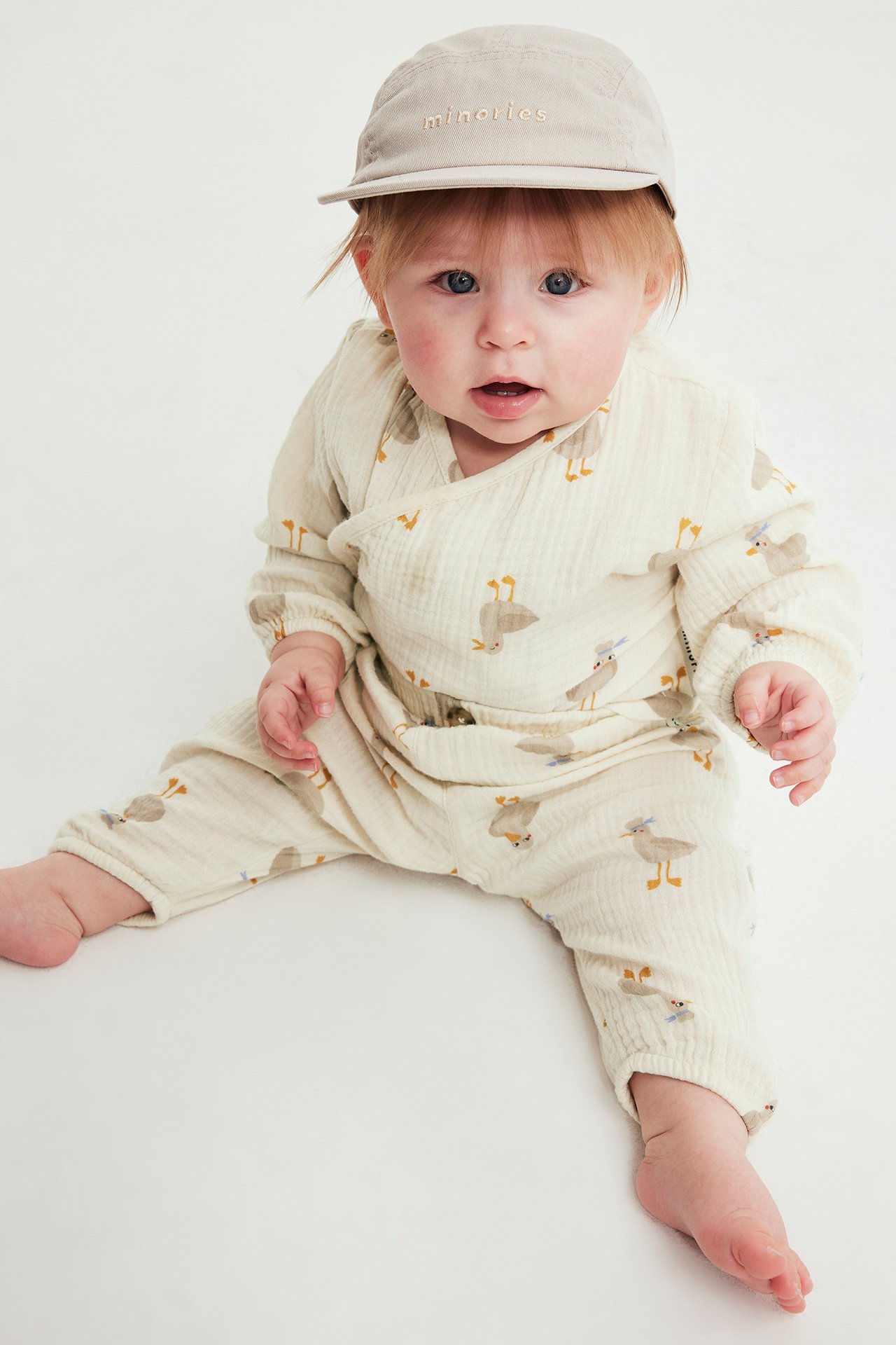 Edestäpäin vauva yllään löysä, beige kuvioitu musliinibody ja beige lippalakki.