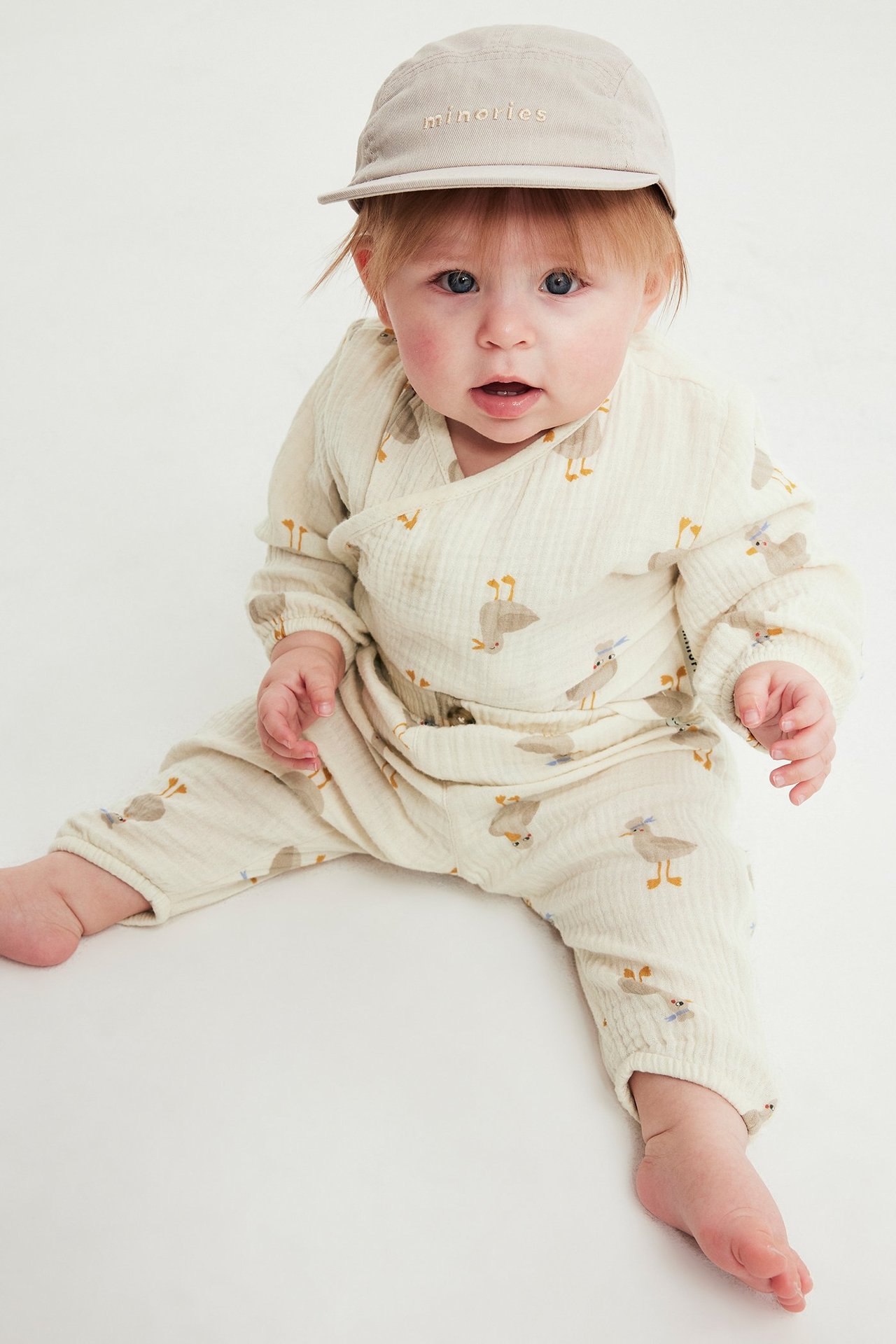 Frontvy av en baby i en lös beige mönstrad muslinbody med omlottknäppning och beige keps.