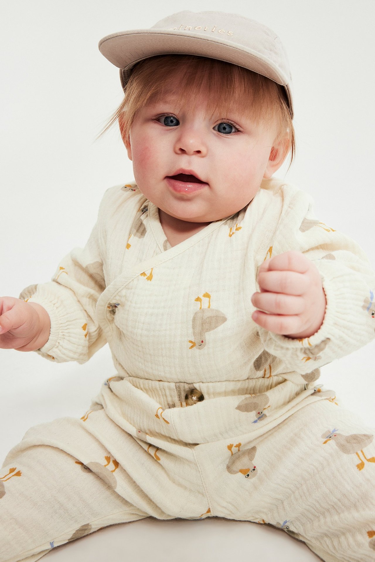 Framifrån, bebis i beige mönstrad långärmad muslinbody med tryckknappar och ljusbeige keps.