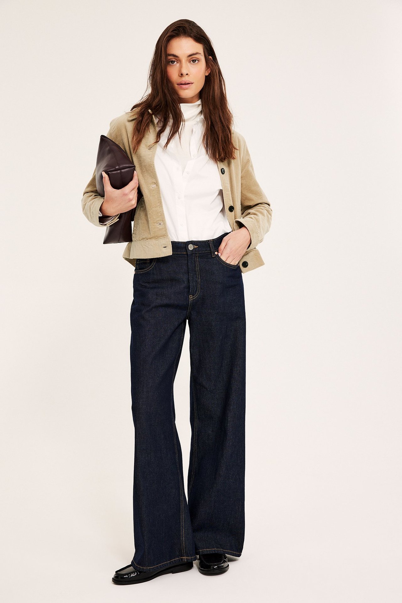 Kvinne i kort beige kordfløyelsjakke, hvit skjorte, mørkeblå jeans og holder en brun clutch.