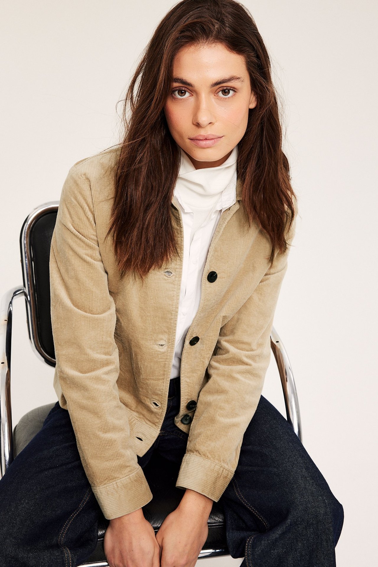 Framifrån, kvinna i beige kort manchesterjacka över vit topp och mörkblå jeans.