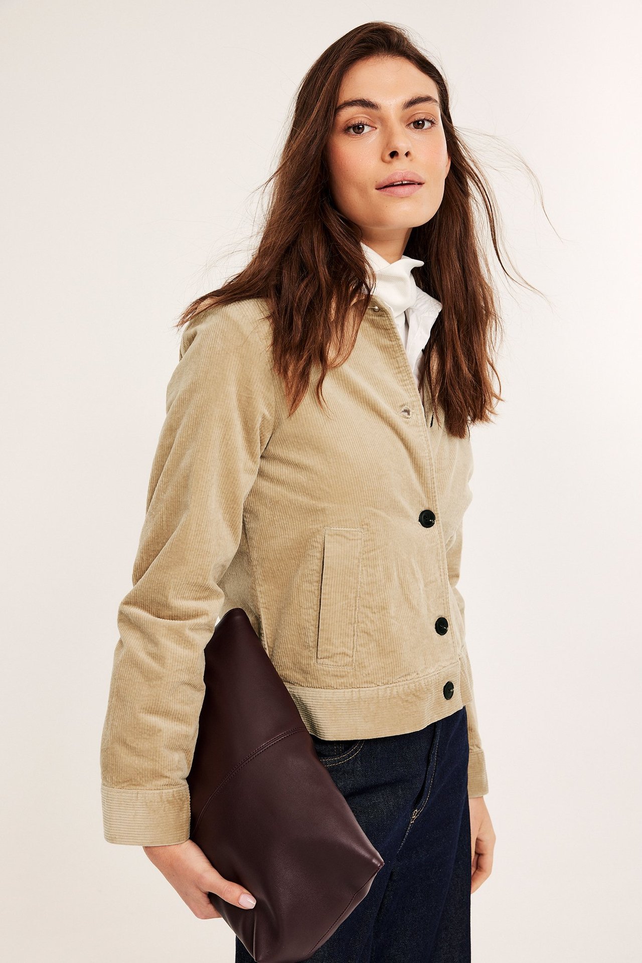 Kvinna i beige semi-kort manchesterjacka, vit topp, mörka jeans, håller en brun clutchväska.