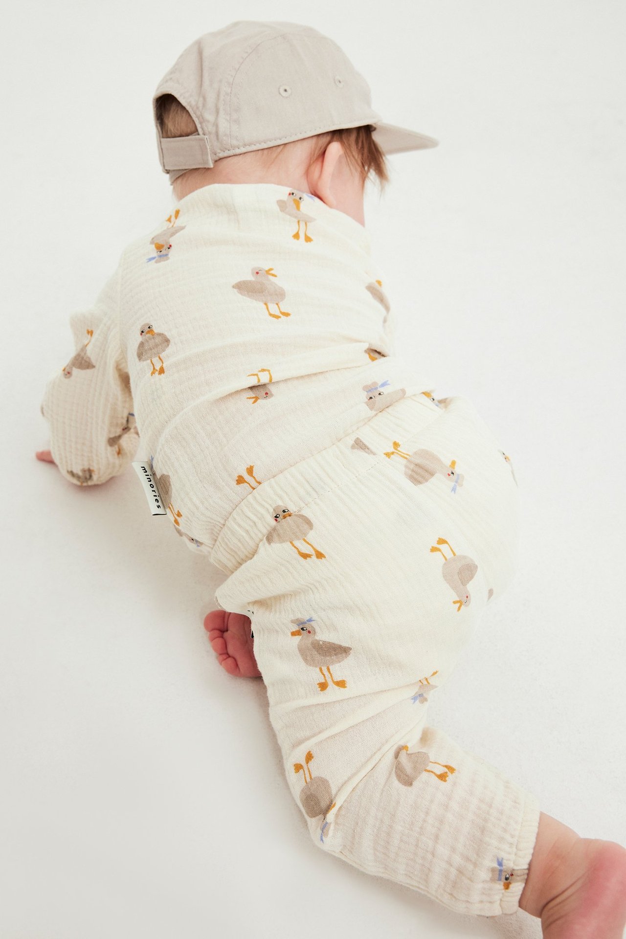 Takakuva ryömivästä vauvasta, yllään beiget kuviolliset musliinihousut, yhteensopiva toppi ja beige lippalakki.