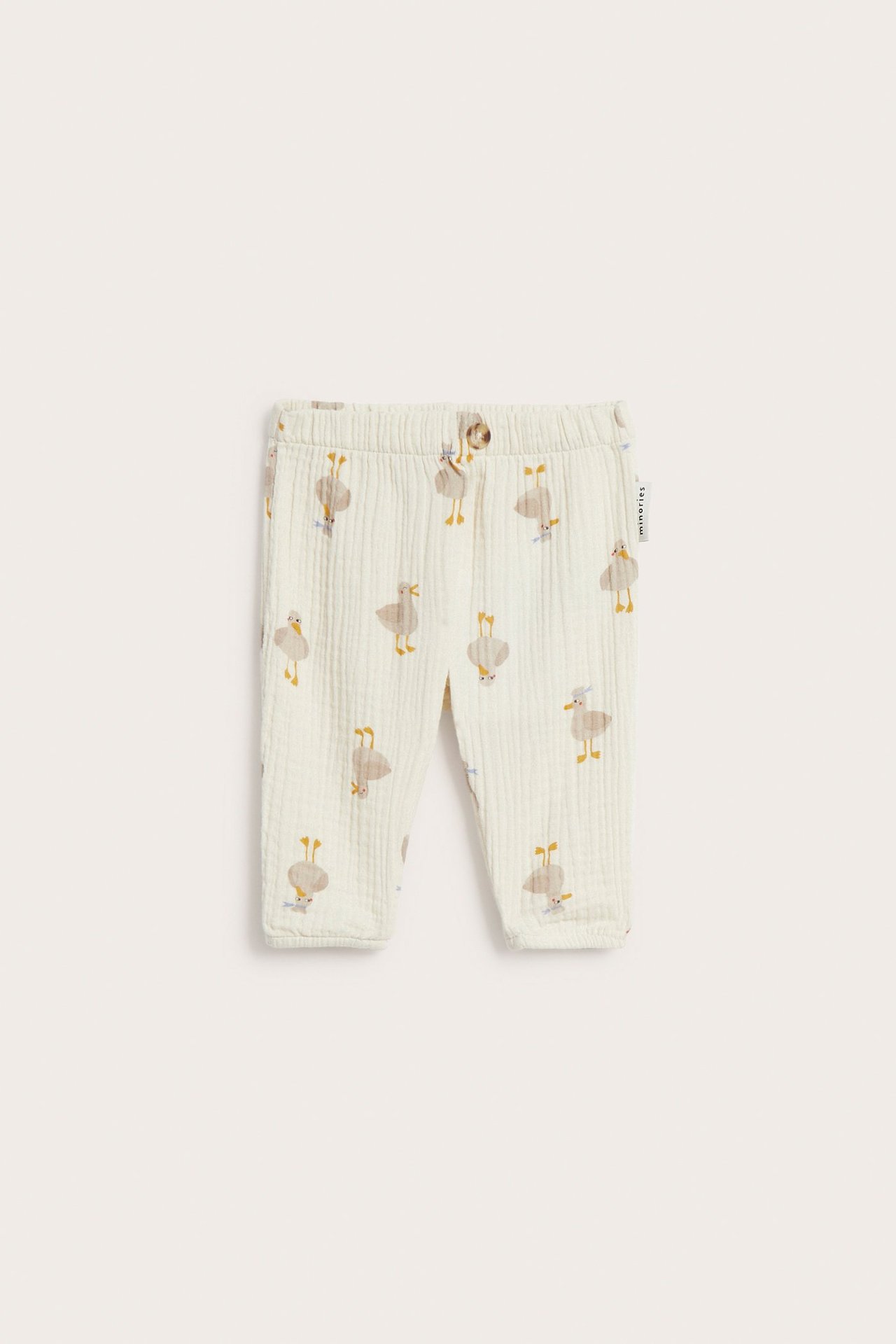 Frontvy av ljusbeige babybyxor i muslin med resår i midjan, fuskknapp och heltäckande fågelmönster.