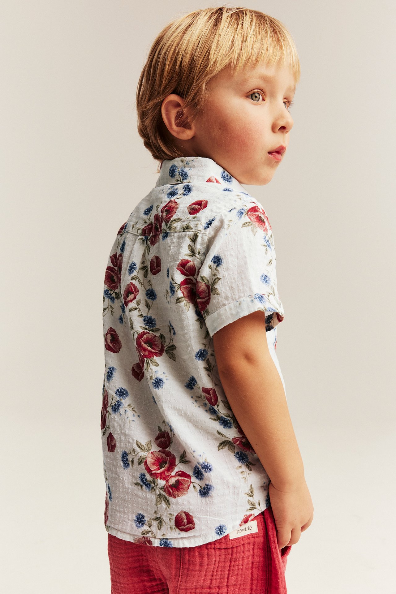 Sidovy av ett barn i en offwhite kortärmad blommig bomullsskjorta och röda texturerade shorts.