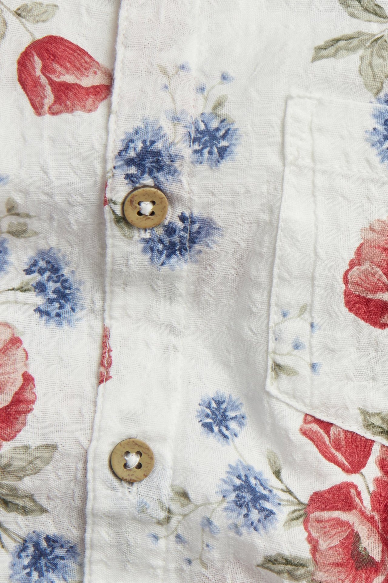 Zbliżenie: kremowa bawełniana koszula, krótki rękaw, czerwono-niebieski kwiatowy wzór, kieszeń i brązowe guziki.