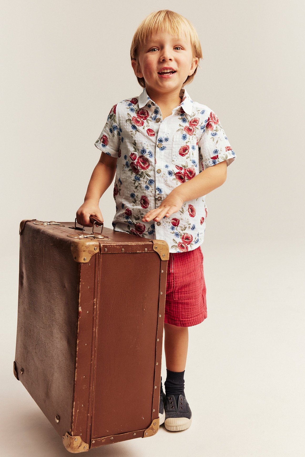 Przodem: Dziecko w krótkiej, kwiecistej koszuli offwhite, czerwonych szortach, trzyma brązową walizkę.