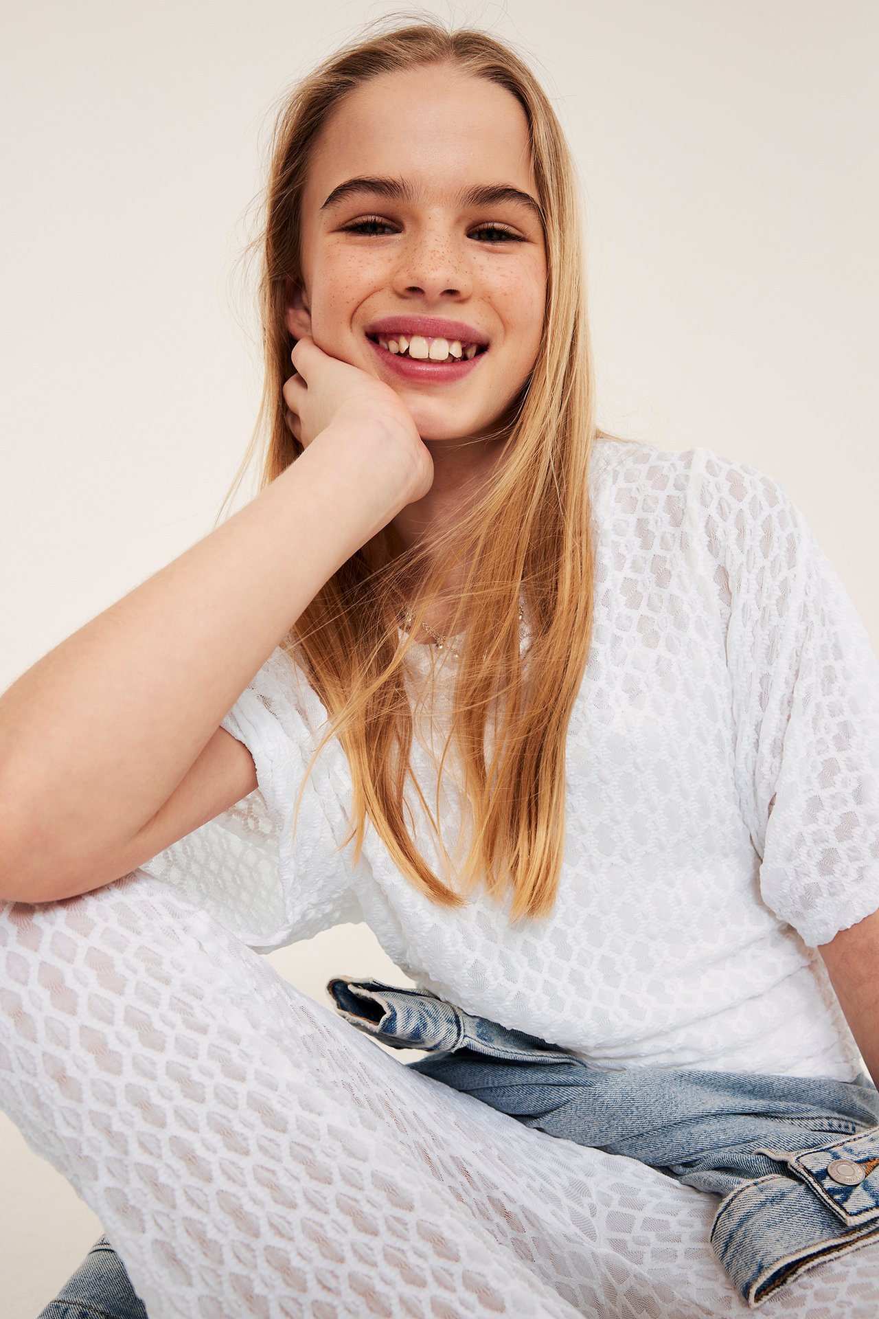 Frontbild på ett leende barn i en vit spetsmönstrad topp med fjärilsärmar och ljusblå jeans.