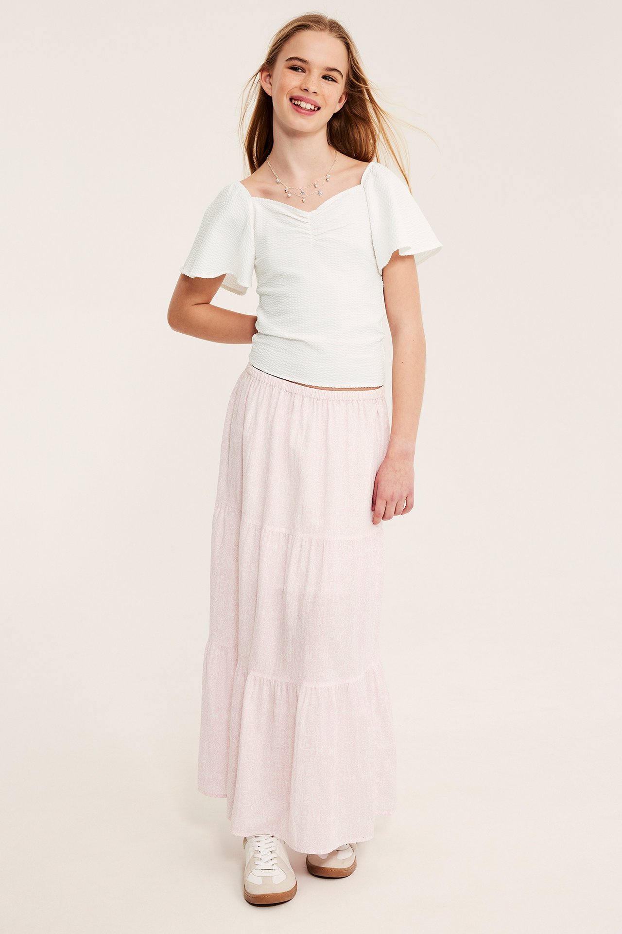 Edestä: Lapsi valkoisessa tiukassa rypytetyssä topissa, perhoshihat, vaaleanpunainen kerroshame ja valkoiset tennarit.