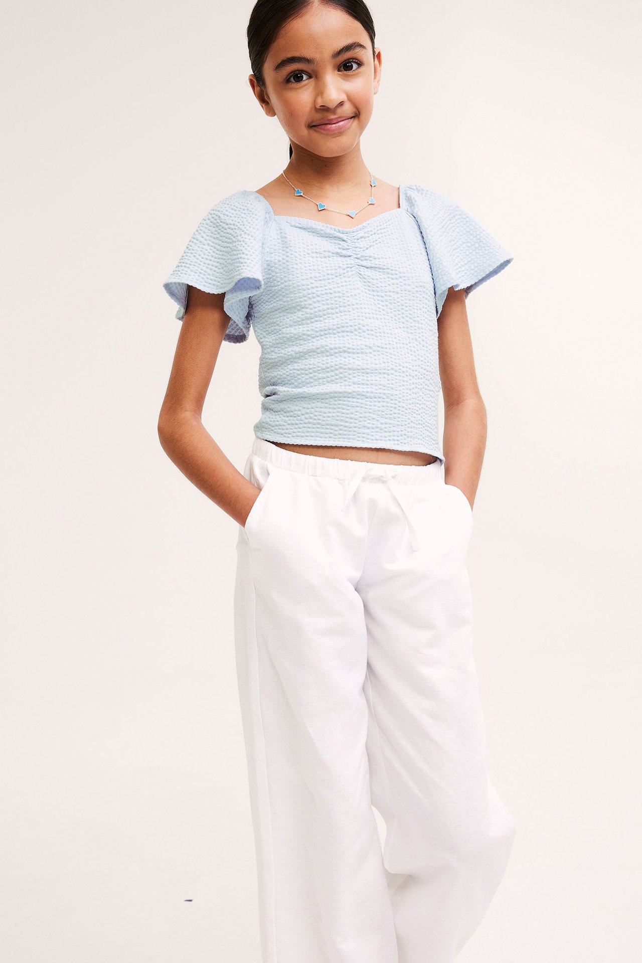 Frontvy: Barn i ljusblå rynkad fjärilsärmad topp, vita vida byxor och ett berlockhalsband.