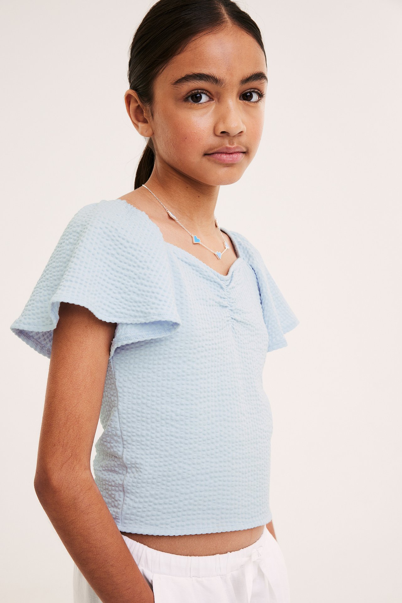 Framifrån: Barn i ljusblå figurnära texturerad topp med rynkad front, fjärilsärm, silverhalsband och vita byxor.