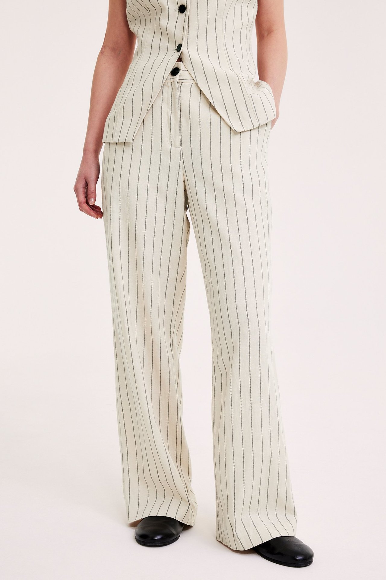 Framsida: Kvinna i offwhite randiga kostymbyxor med vida ben och matchande randig väst med svarta knappar.