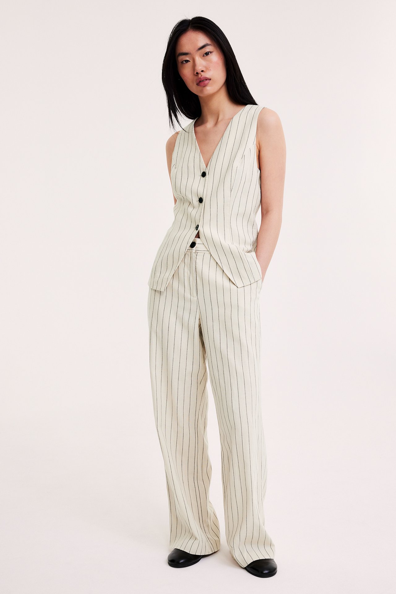 Widok z przodu: kobieta w kremowych, pasiastych lnianych spodniach z szerokimi nogawkami i pasującej kamizelce.