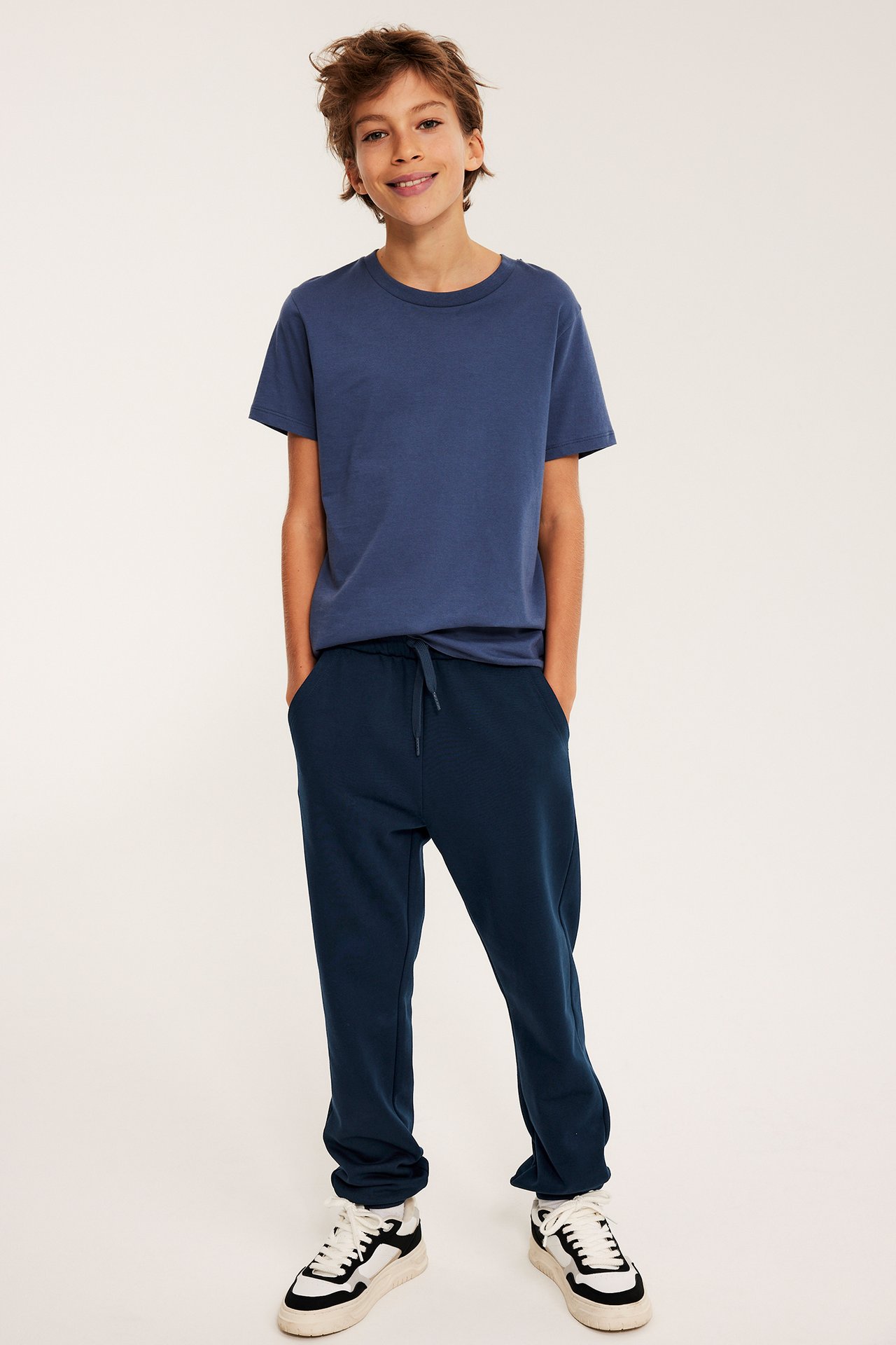 Widok z przodu: dziecko w ciemnoniebieskich spodniach dresowych ze sznurkiem, niebieskiej koszulce i białych sneakersach.