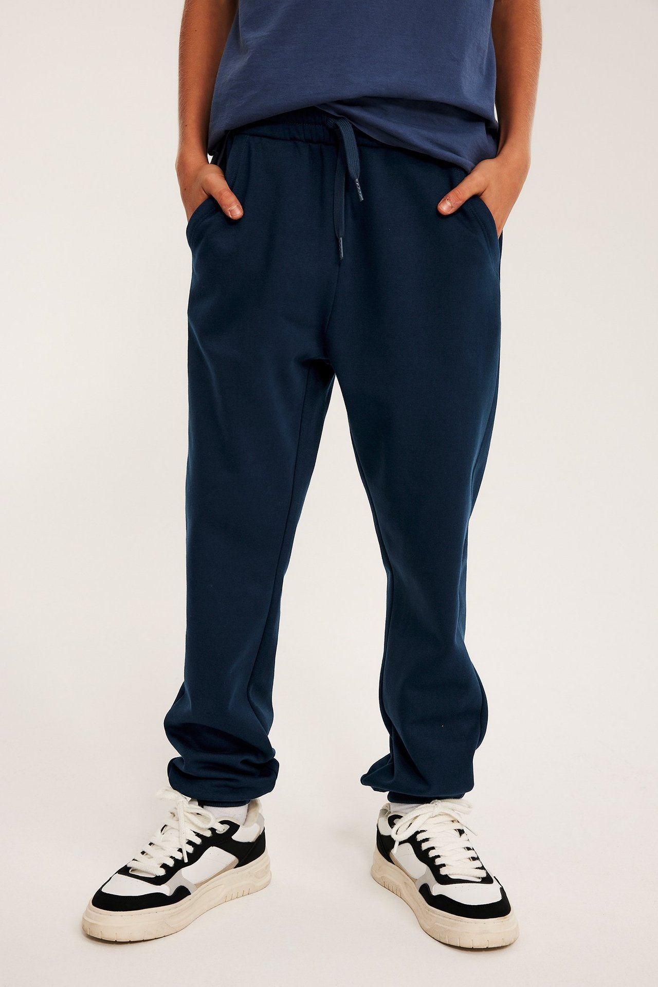 Widok z przodu: Dziecko w granatowych dresach (sznurek, kieszenie), granatowej koszulce, białych trampkach.