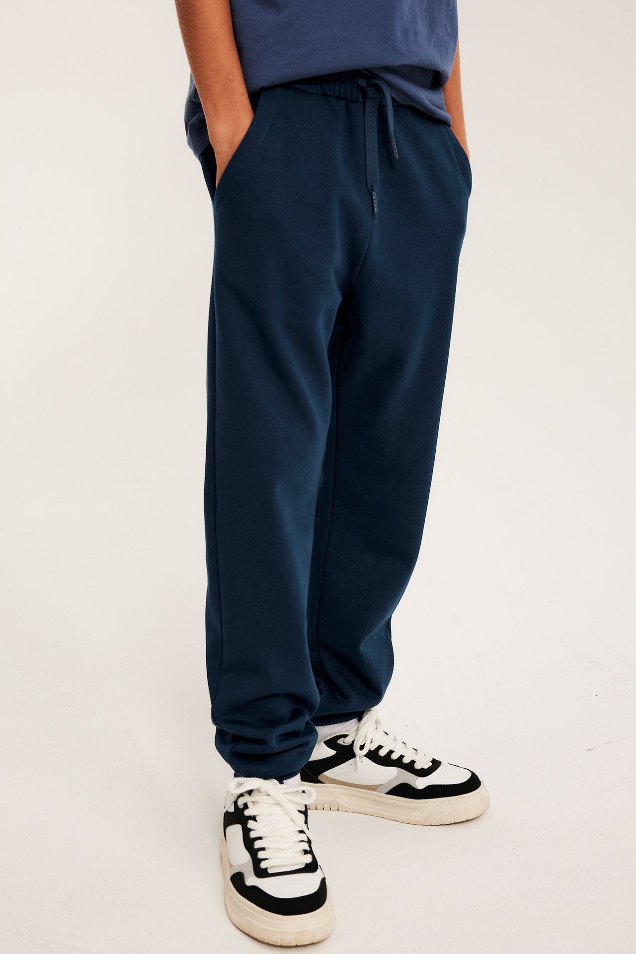 Widok z przodu: dziecko w granatowych dresach z rękami w kieszeniach, niebieskiej koszulce i biało-czarnych sneakersach.