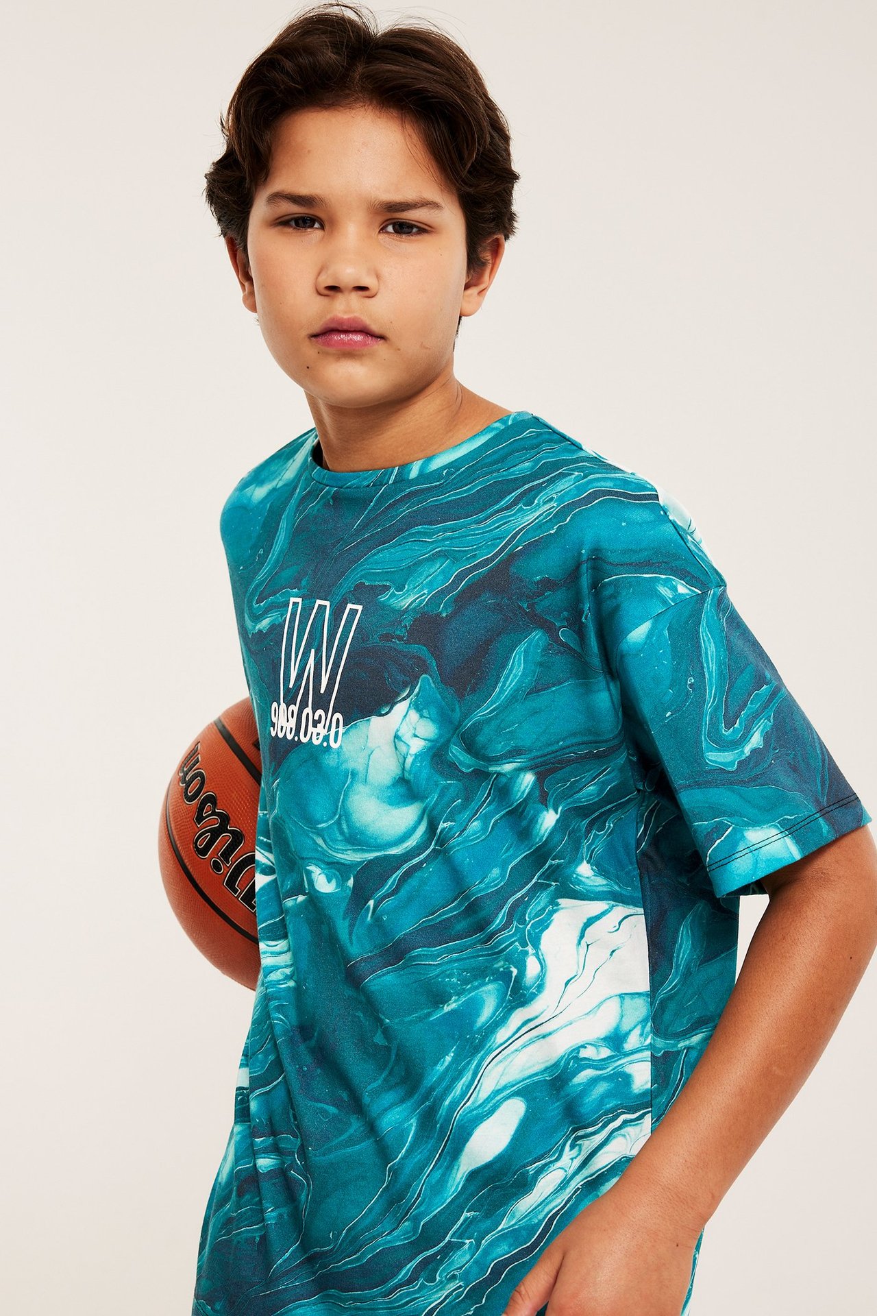 Edestäpäin: Lapsi yllään ylisuuri turkoosin ja valkoisen marmoroitu t-paita graafisella painatuksella, pitäen oranssia koripalloa.