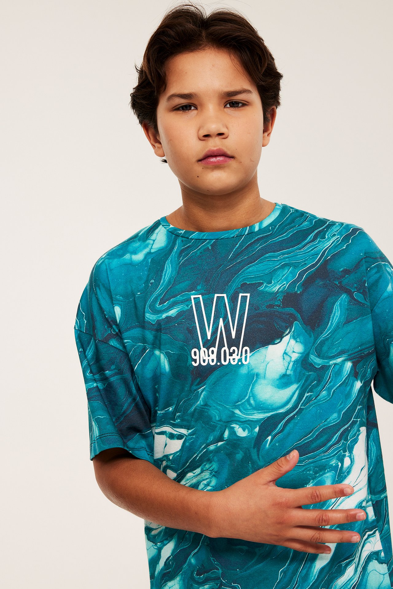 Edestä näkymä lapsesta, yllään väljä turkoosi-valkoinen marmoroitu t-paita valkoisella kuvalla.