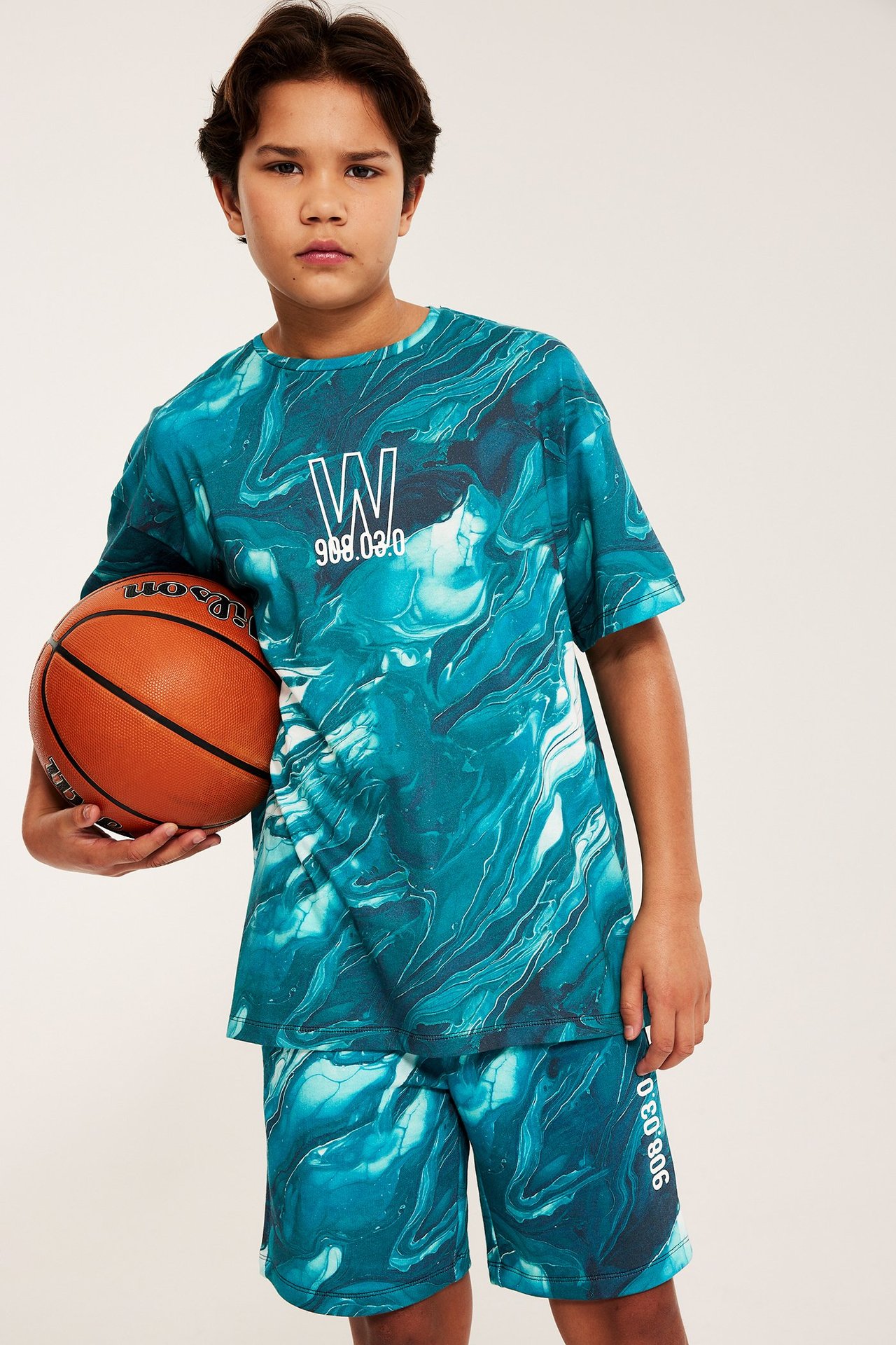 Widok z przodu dziecka w luźnym turkusowym t-shircie z wirującym wzorem i pasujących szortach, trzymającego piłkę do koszykówki.