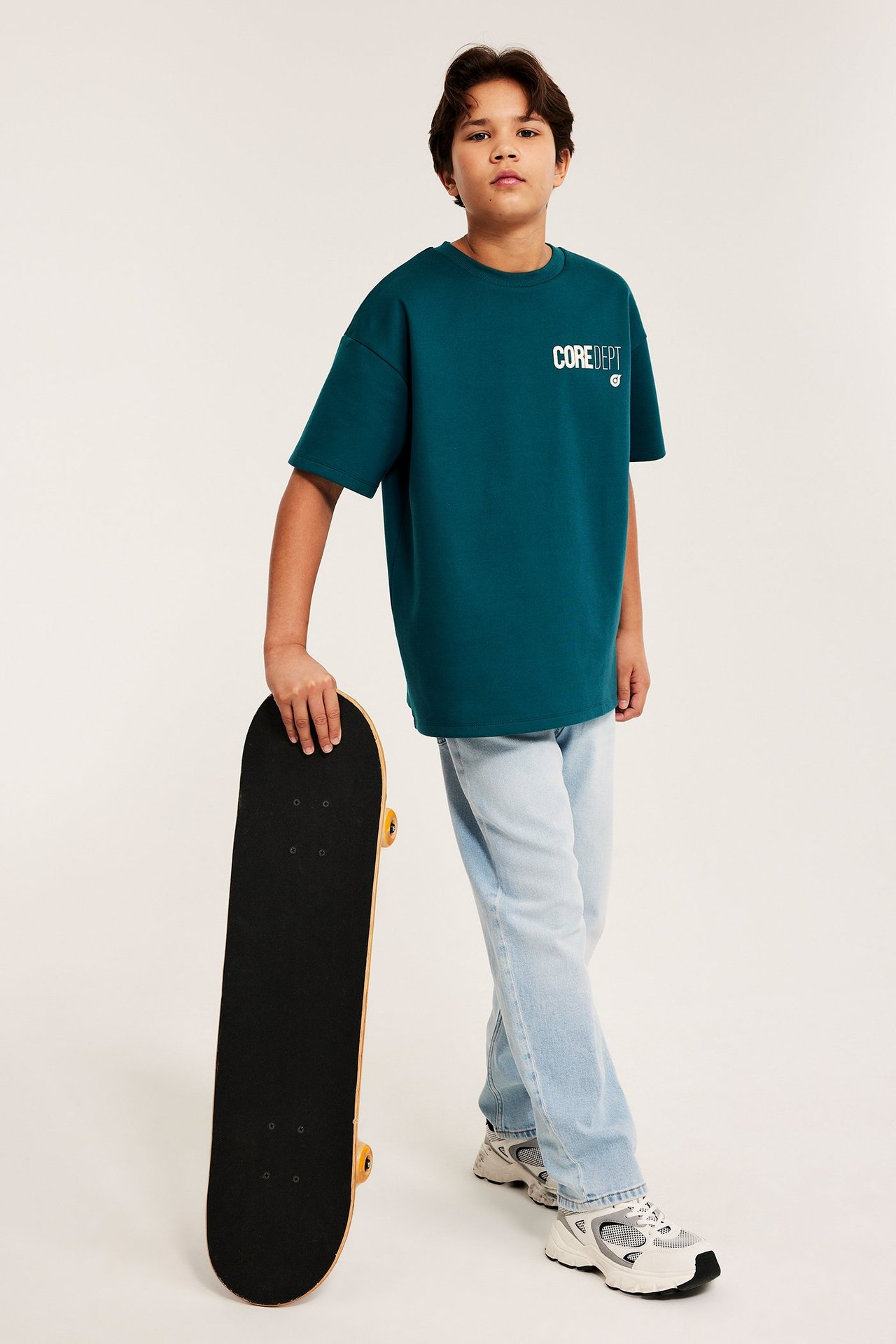 Lapsi vihreässä ylisuuressa printtipaidassa, vaaleansinisissä farkuissa, valkoisissa lenkkareissa, pitäen skeittilautaa.