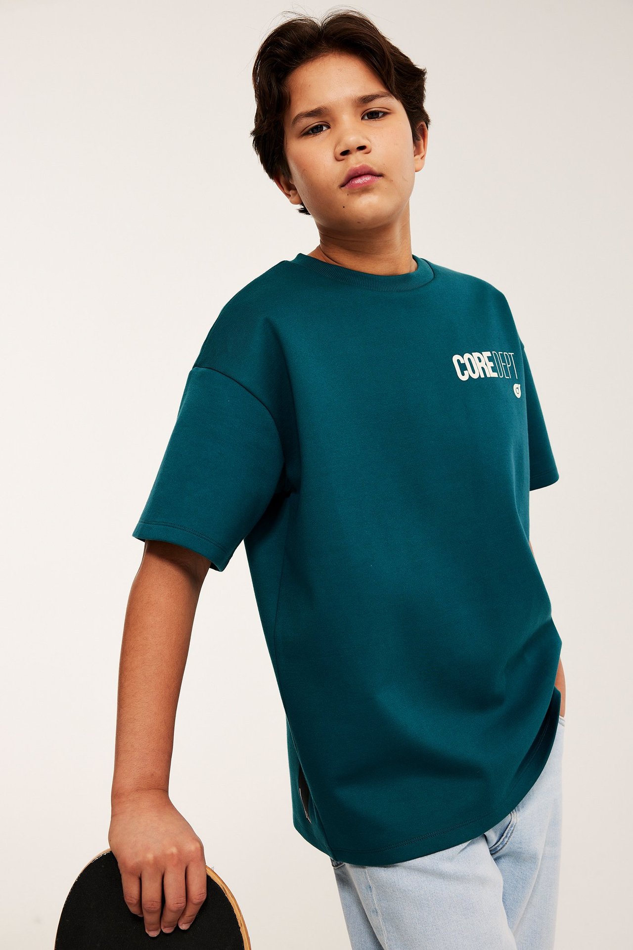 Edestä: Lapsi vihreässä oversize-t-paidassa, jossa valkoinen painatus, vaaleat farkut, pitää skeittilautaa.