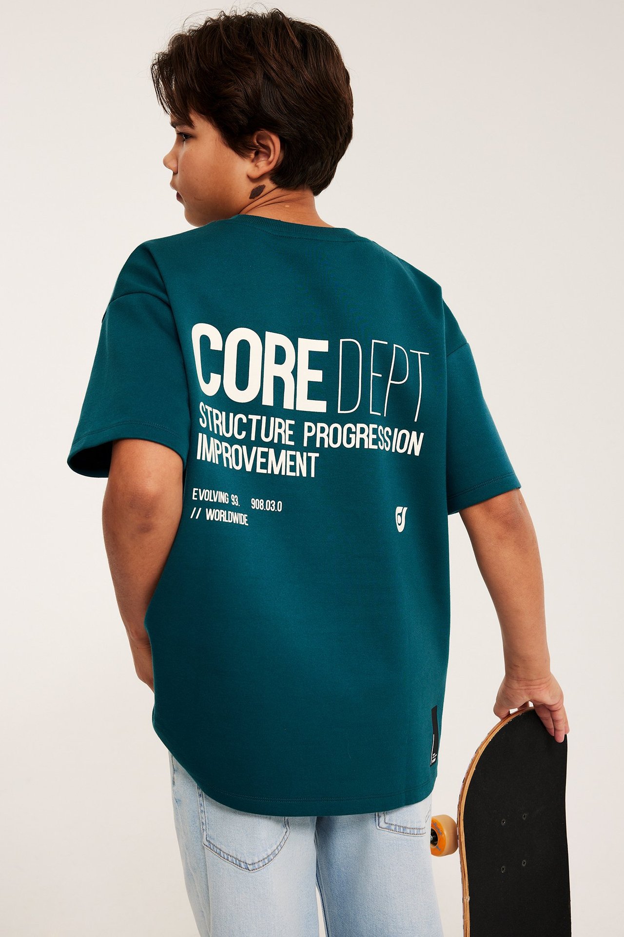 Widok z tyłu dziecka: zielona koszulka oversize z białym nadrukiem, jasnoniebieskie jeansy, deskorolka.