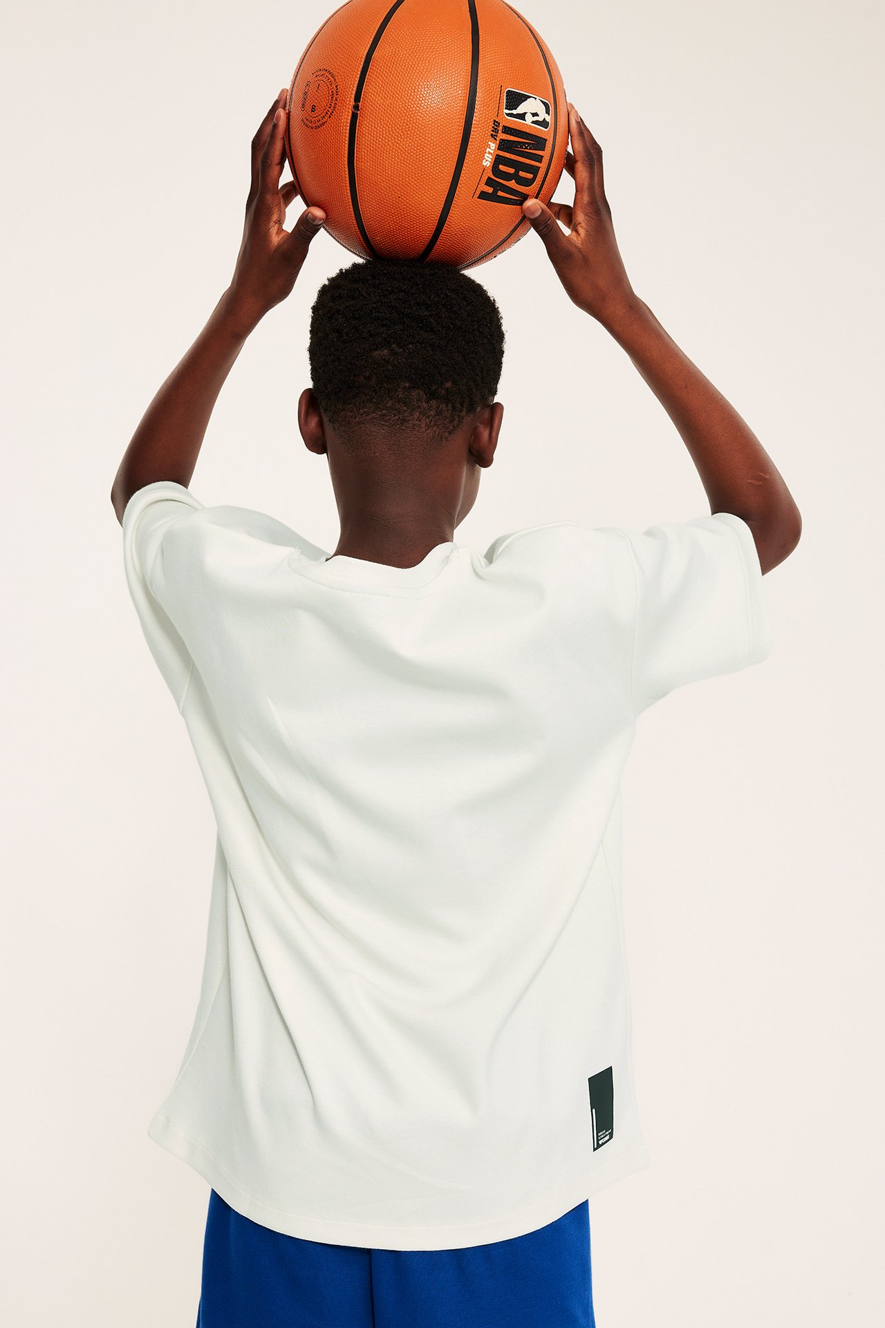 Takakuva lapsesta valkoisessa oversized T-paidassa ja sinisissä shortseissa, pitelee koripalloa.