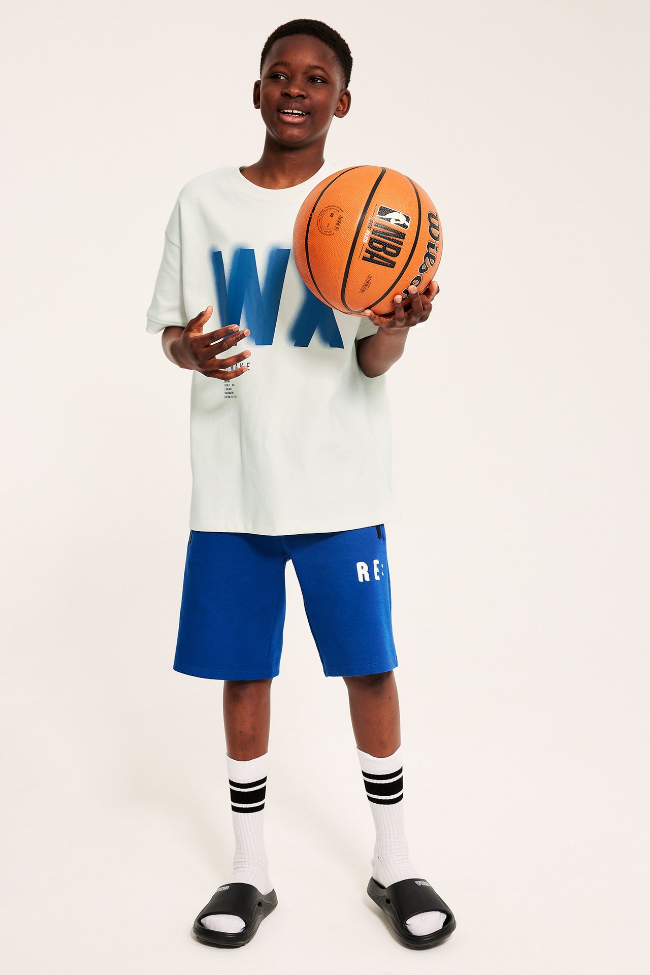 Forfra: Barn i hvit oversized t-skjorte med 'WX'-trykk, blå shorts, stripete sokker, sorte sandaler, holder basketball.
