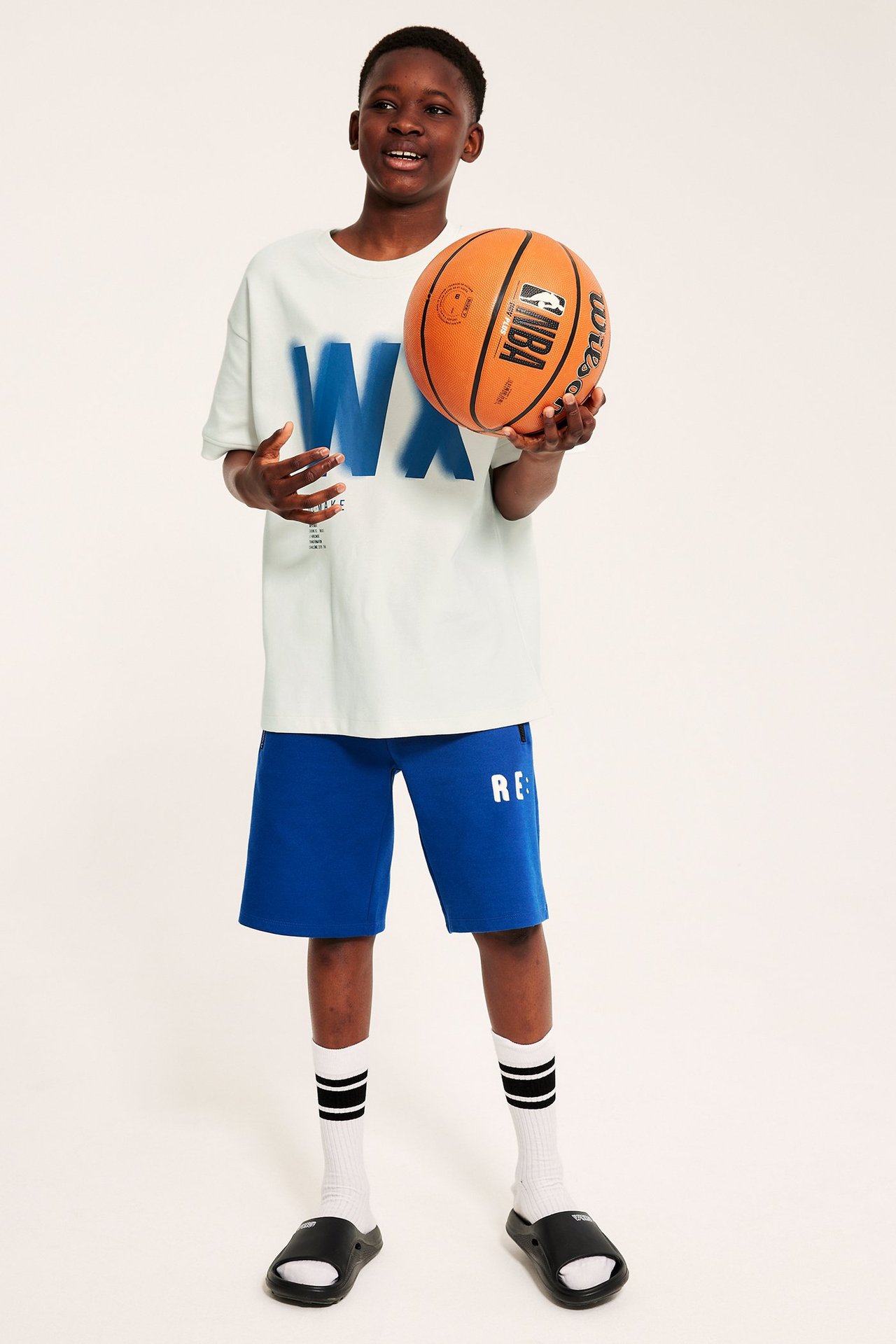 Framifrån: Barn i vit oversized t-shirt med 'WX'-tryck, blå shorts, randiga strumpor, svarta tofflor, håller basketboll.