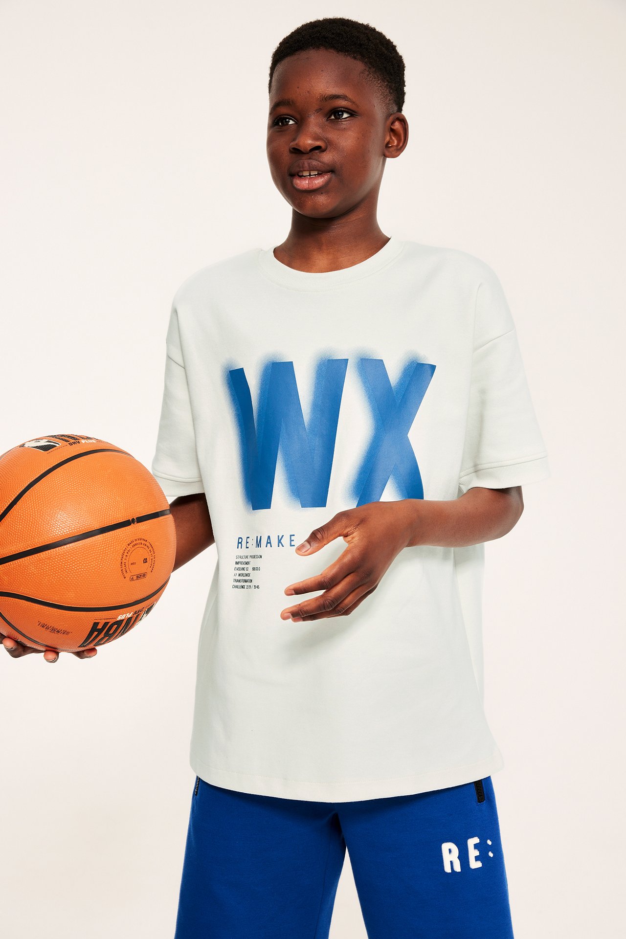 Frontvy av ett barn i vit oversized t-shirt med blått tryck, blå shorts, håller en basketboll.
