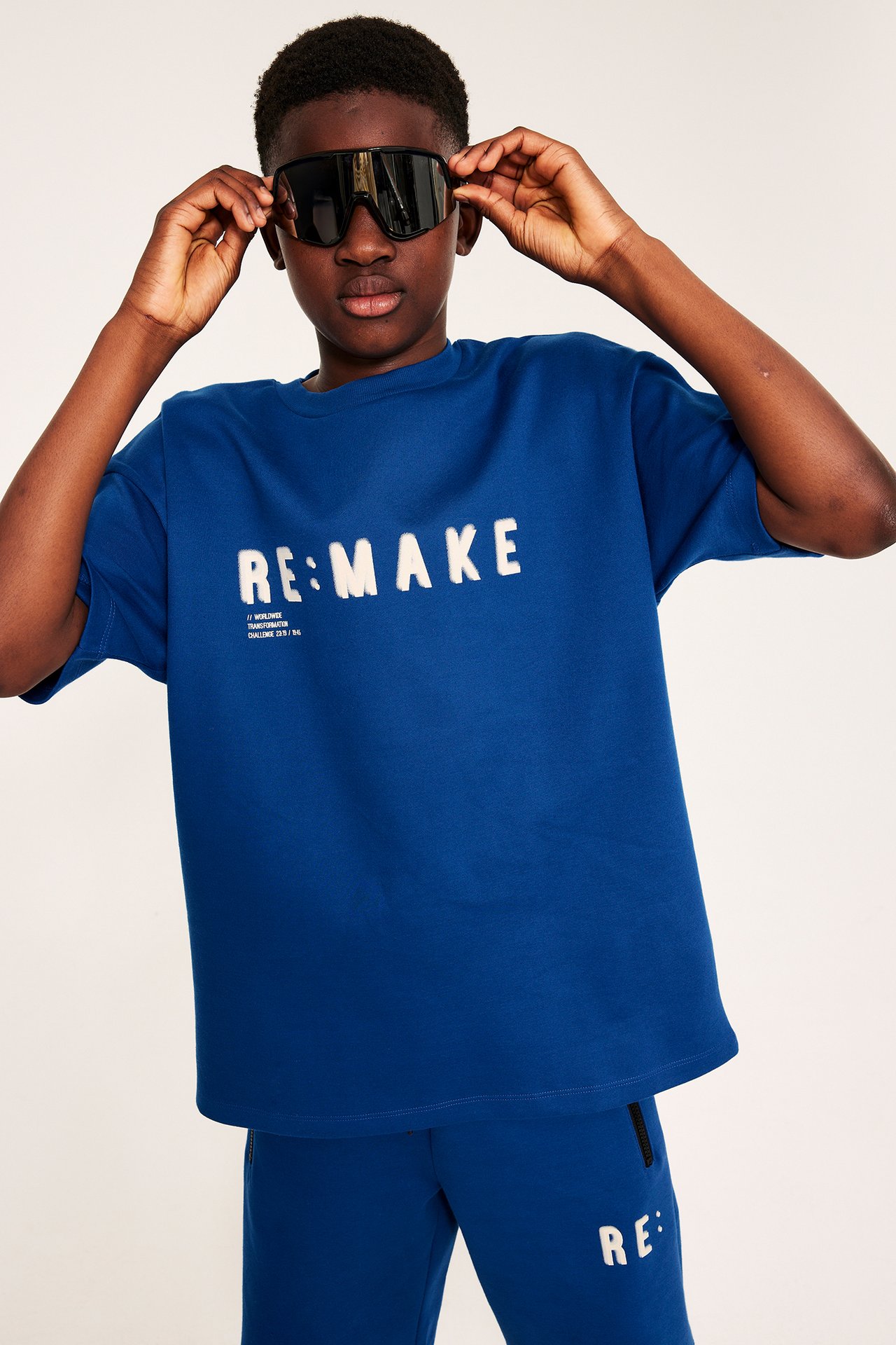 Widok z przodu: Dziecko w granatowym, oversize’owym t-shircie „RE:MAKE” i pasujących dresach, poprawiające czarne okulary.